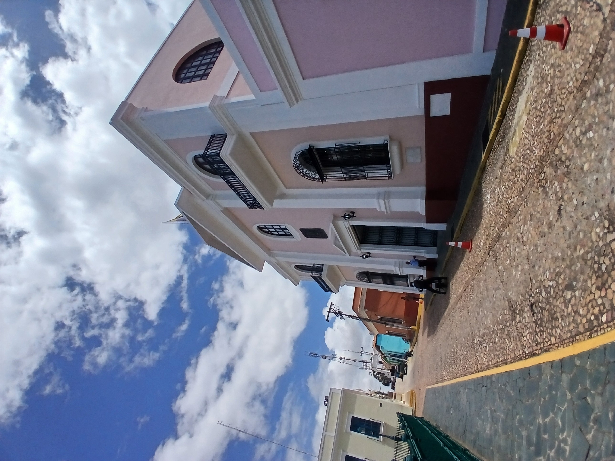 The image depicts a street scene in Unknown, Unknown, showing a view of a building and the sky. The building is primarily pink and white, with architectural details such as arched windows and a balcony. The base of the building is maroon. On the street, two traffic cones are visible, one partially cut off in the top right corner, with alternating red and white stripes.
People can be seen on the street, with one individual appearing to be walking. The street itself is paved with cobblestones and is bordered by a yellow line. In the background, there are power lines and additional buildings of different colors. The sky above is blue with white clouds.