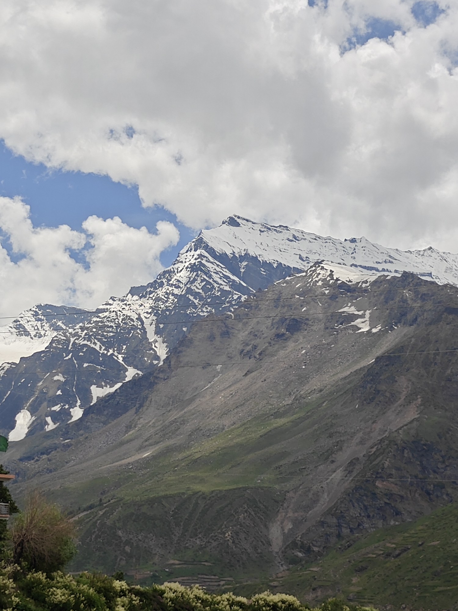 The image captures a majestic snow-capped mountain range under a partly cloudy sky. The mountains are rugged, with patches of snow clinging to their peaks and slopes, interspersed with exposed rock and scree. Lush green vegetation covers the lower slopes, giving way to a more arid, rocky terrain higher up. In the foreground, a few trees and bushes are visible, with some white flowering plants at the very bottom. A few power lines are also present, stretching horizontally across the mountain face. The overall scene suggests a natural landscape in a mountainous region, likely a tourist destination given the presence of what appears to be some sort of building with a green flag in the lower left corner, possibly indicative of a viewpoint or a guesthouse. The location is identified as Yamling, India, a place known for its scenic beauty and mountainous terrain.