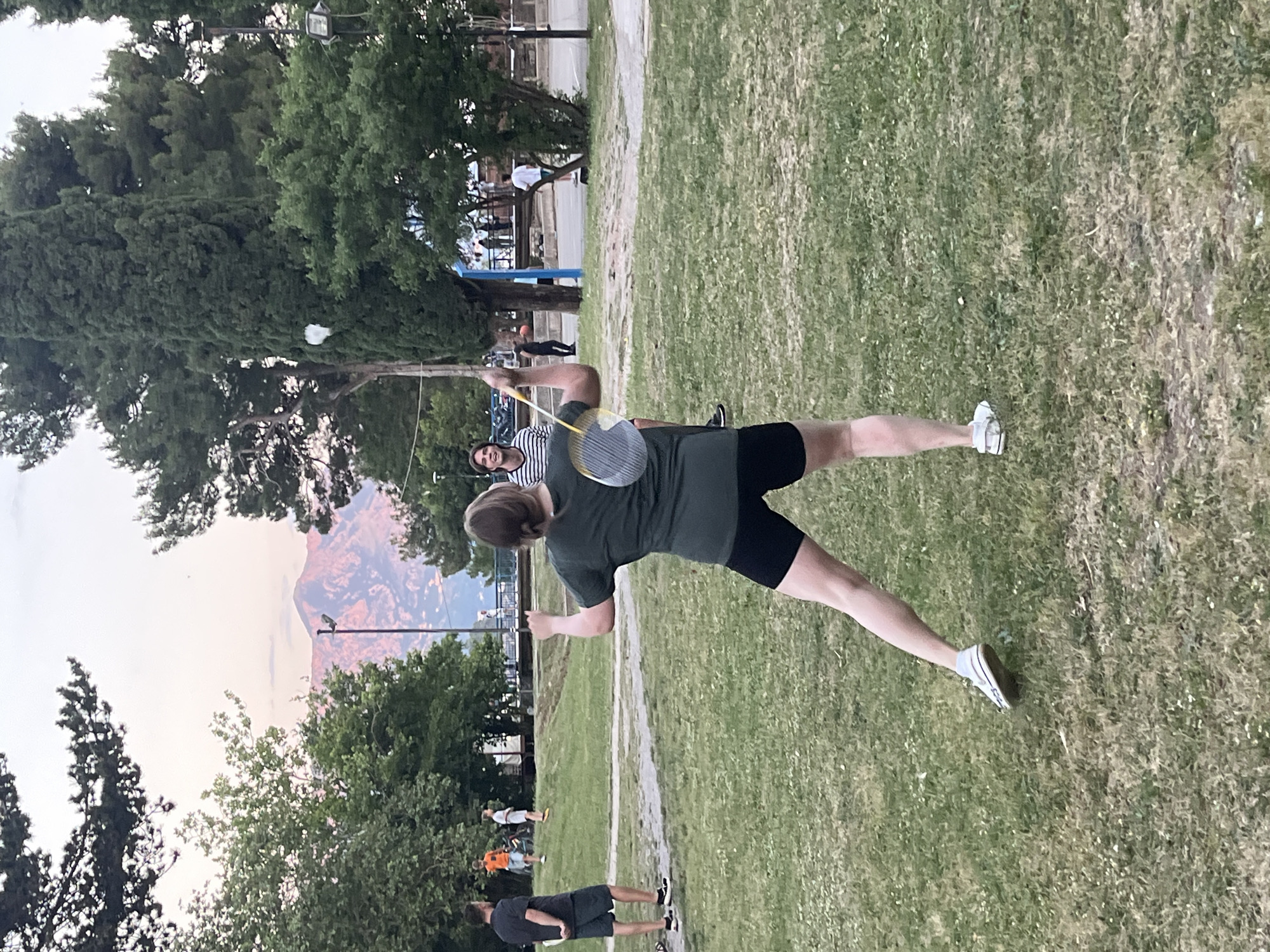 The image depicts a person playing badminton outdoors in Budva, Montenegro. The person is captured in mid-swing, holding a badminton racket, with a shuttlecock visible in the air. They are wearing a dark-colored t-shirt and shorts, and white sneakers. A second person stands nearby, possibly watching the game. Several other people are situated in the background, along with trees and a mountain range in the distance. The scene is set on a grassy area, likely a park or recreational space. The image has been rotated, but the details of the location and activities remain clear.