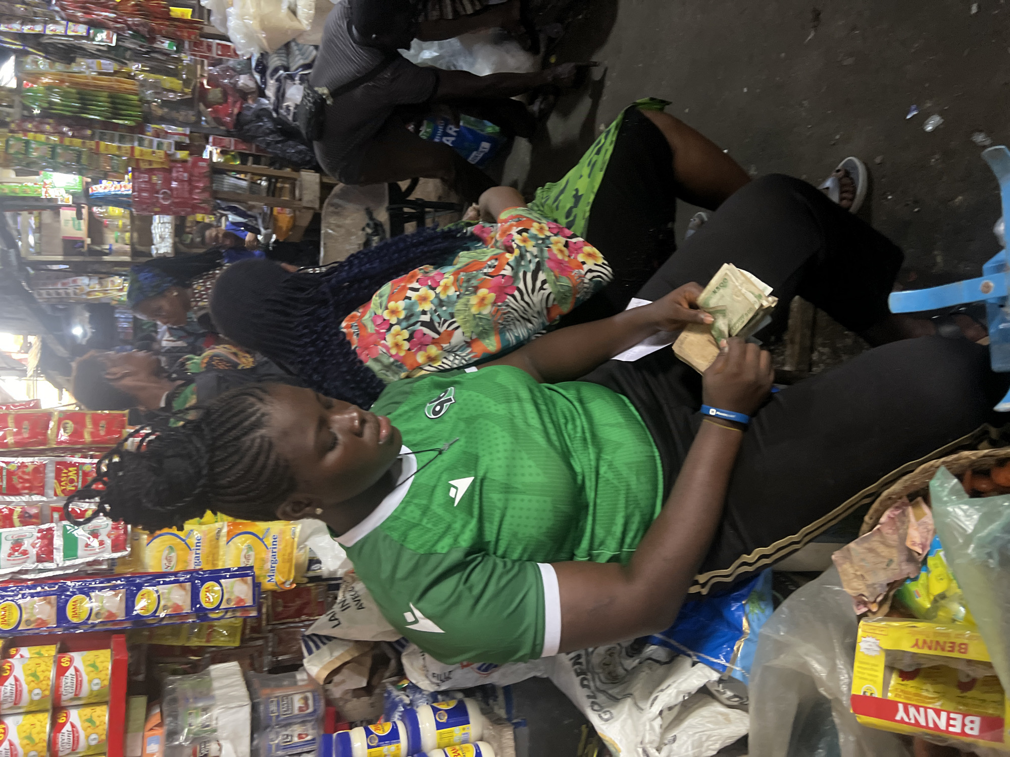 In Nkpor, Nigeria, a young woman wearing a green soccer jersey with white accents and black athletic pants is seated and counting money. Her hair is styled in braids that are pulled up into a bun. She is holding several Nigerian Naira banknotes. The setting appears to be a market stall, with shelves stocked with various goods, including canned food, packets of spices or seasonings, and other packaged items, visible in the background. Other individuals are present in the scene, including a woman with a colorful floral top and dark braided hair seated next to her, and other people blurred in the background, suggesting a busy marketplace environment. The ground is dark and appears to be paved or compacted earth, with some scattered debris.