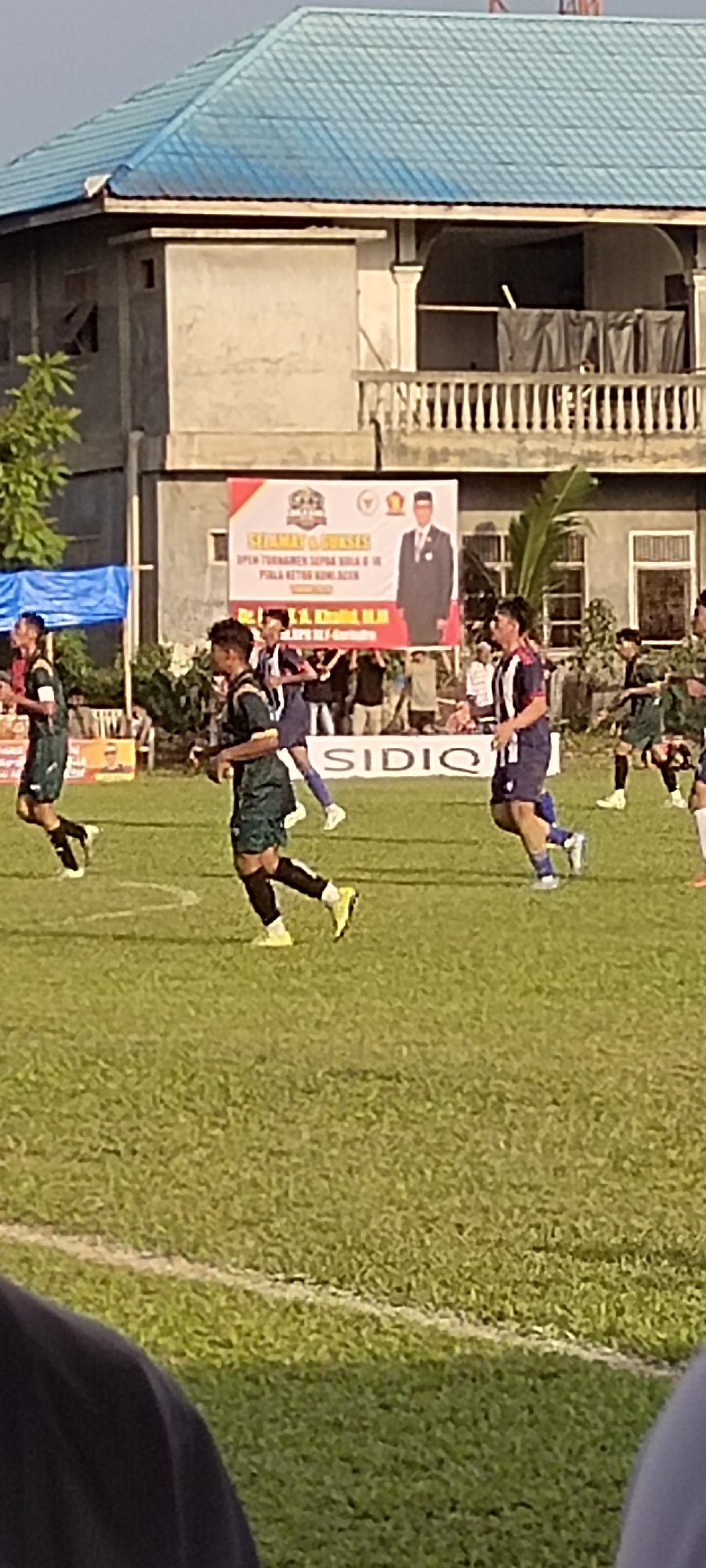The image depicts a soccer match taking place on a grassy field in Keudesimpangempat, Indonesia. In the background, a building with a blue tiled roof is visible, along with a banner advertising an event. Several soccer players in green and blue uniforms are on the field, engaged in the game. Some players are running, while others appear to be observing the play. The lighting suggests it is daytime, possibly late afternoon. The environment is an outdoor sports venue.
