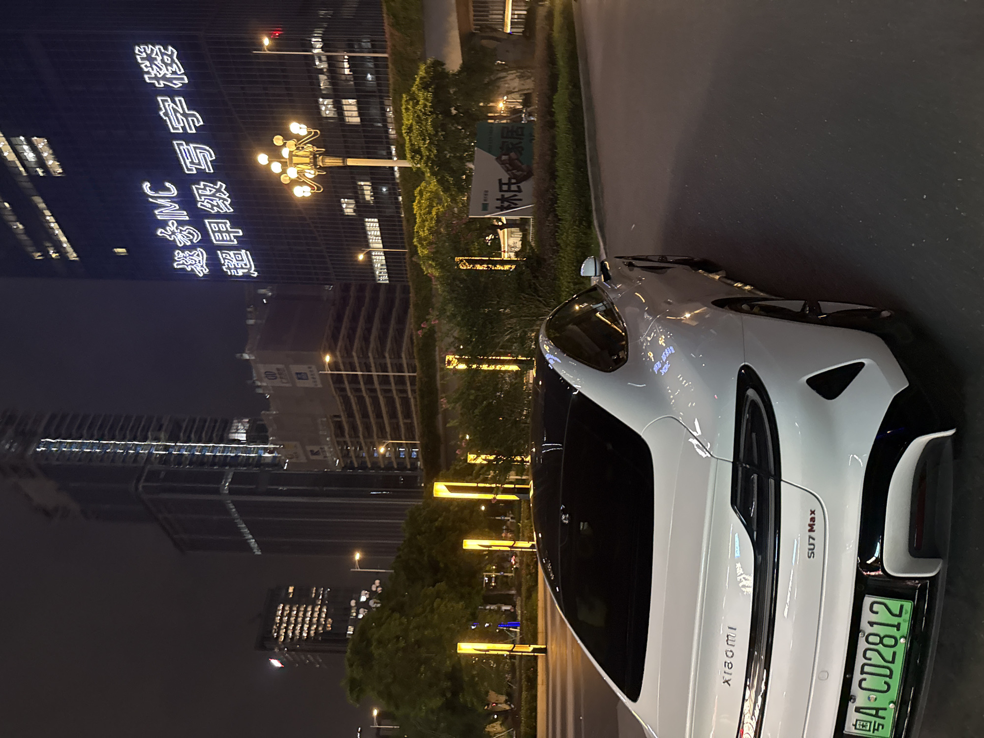 The image shows a white Xiaomi SU7 Max car parked on a street at night in Huangpu District, China. The car is positioned on the right side of the frame, with its rear facing the camera. The background features illuminated skyscrapers, one of which has large white characters displayed on its facade. A street lamp with multiple bulbs is visible in front of the illuminated building. To the left of the car, there are several tall, vertical yellow lights and trees. A sign with Chinese characters is also visible in the middle-ground, to the right of the car, depicting what appears to be a company logo and text. The street surface is asphalt.