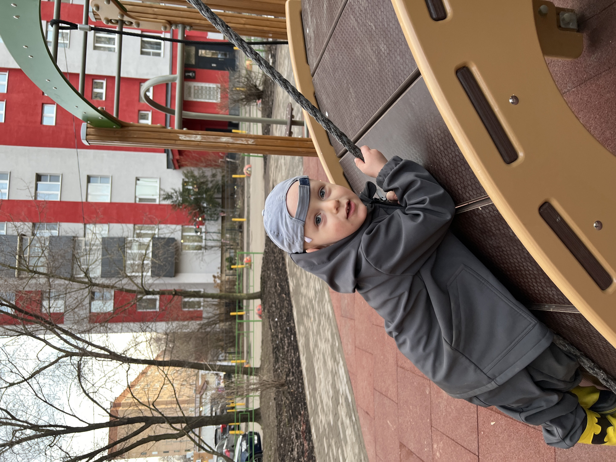 A young child, wearing a gray hooded jacket, pants, and a baseball cap, is on playground equipment. The playground is located in Mytishchi, Russia. The child is gripping a dark-colored rope attached to the playground structure. The playground surface is red-brown. In the background, there is a multi-story building with red and white paint. There are also trees, some bare and some with minimal foliage, as well as street furniture and parked cars visible.