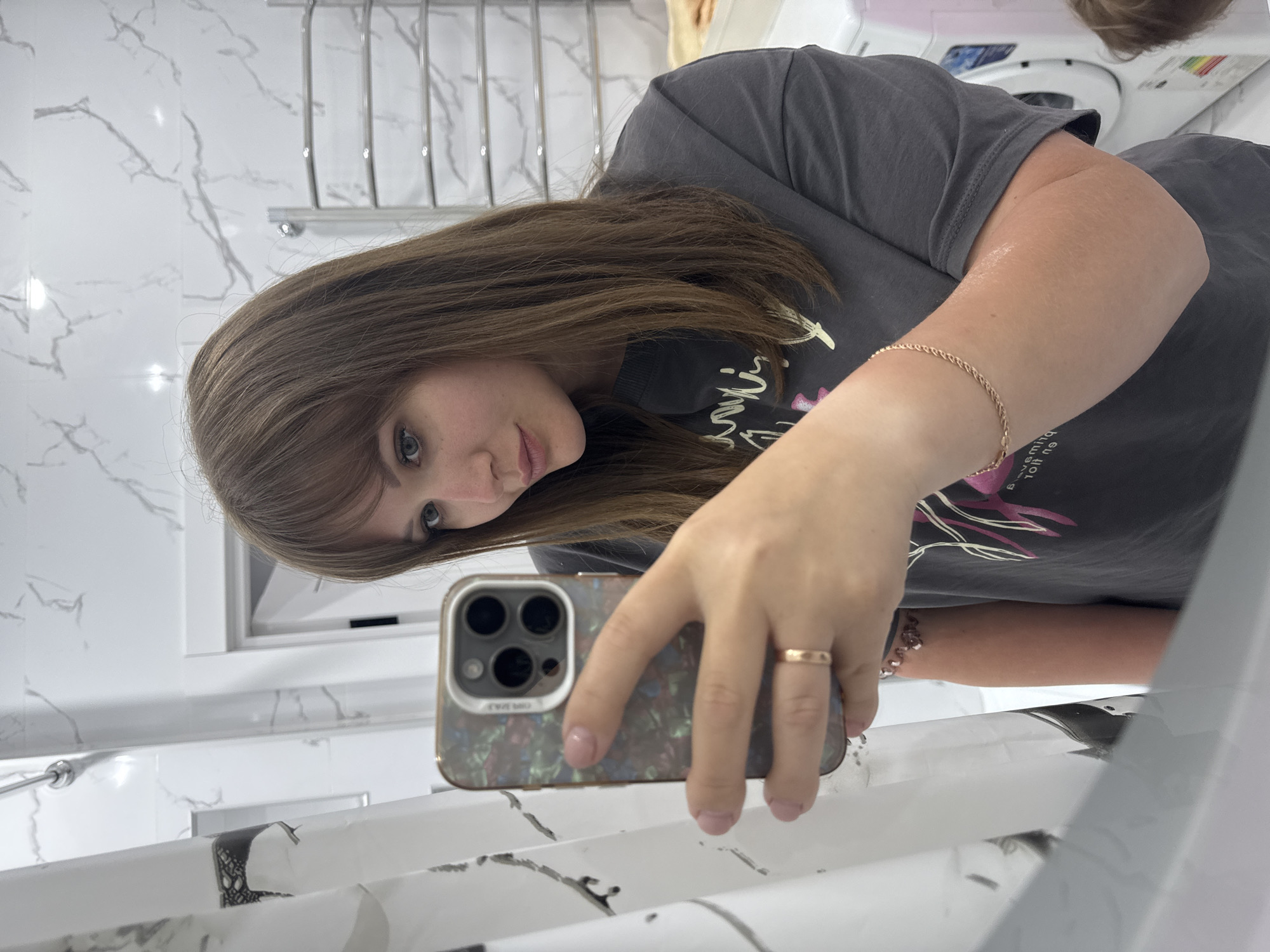 A woman is taking a selfie in what appears to be a bathroom. She is holding a smartphone with a multi-camera setup. She is wearing a gray T-shirt and has a gold ring on her finger and a gold bracelet. The background shows a marble-tiled wall, a towel rack, and a washing machine. While the location cannot be confirmed, the prompt asked if it could be Stavropol, Russia. The information to confirm or deny that location is not contained within this image.