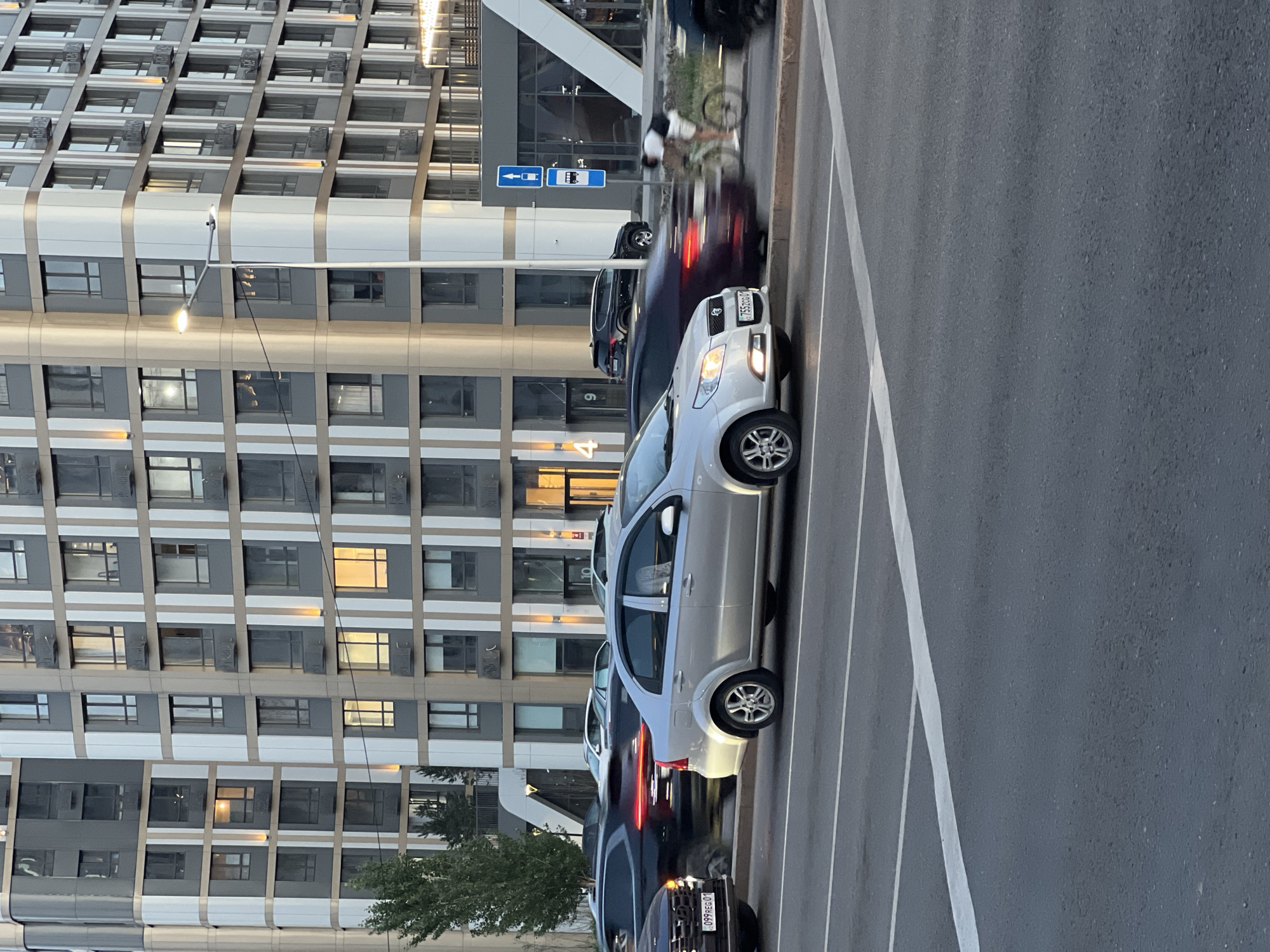 The image captures a street scene in Astana, Kazakhstan, with a modern residential building dominating the background. The building features a distinctive facade of grey and white panels with numerous windows, some illuminated, suggesting residential occupancy. A street lamp is visible, casting a warm glow on the building's corner. In the foreground, several cars are in motion on a multi-lane road. A silver sedan is prominently featured in the center, with its headlights on. Other vehicles, including a dark SUV and a dark sedan, are also visible, some with motion blur indicating their movement. A person on a bicycle is also present, moving in the same direction as the traffic. Traffic signs, including a bus stop sign, are posted on the side of the road. The road is marked with white lane lines, and the overall impression is of an urban environment with active traffic.