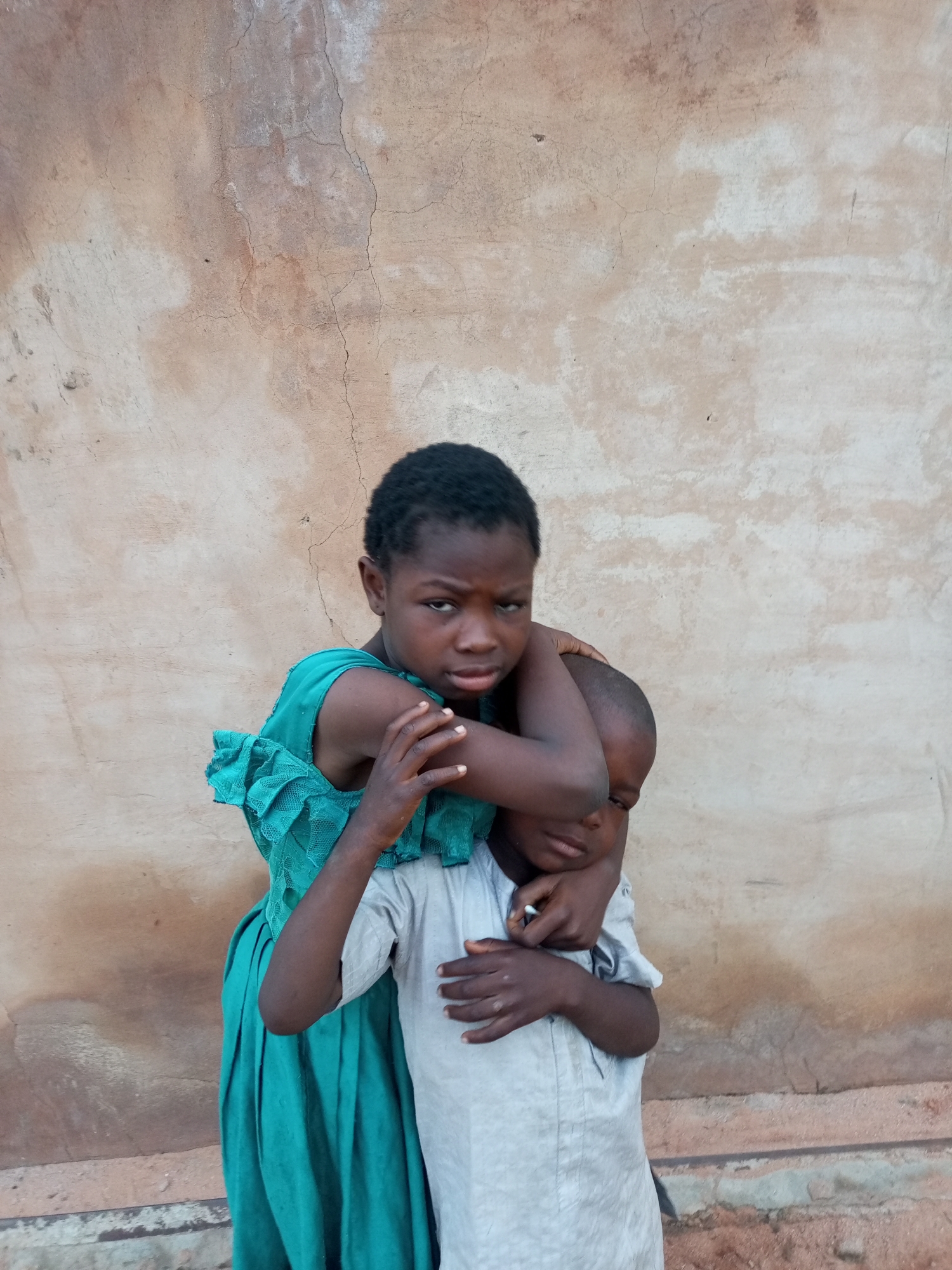 Two children are standing in front of a rough, textured wall in what appears to be an outdoor setting in Tempure, Nigeria. The child in the foreground, on the left, is wearing a teal dress with ruffled sleeves and is embracing the child behind them. This child has dark skin, short black hair, and is looking directly at the camera with a serious expression. Their left arm is wrapped around the neck of the other child.
The second child, positioned behind and slightly to the right of the first, is wearing a light gray, long-sleeved garment. Only their head and upper torso are visible. Their face is partially obscured by the arm of the child in front, and they are looking downwards. Their hands are clasped together in front of their chest.
The wall behind the children is light brown with patches of lighter and darker discoloration, and appears weathered with visible cracks and imperfections. The ground in front of the wall is not fully visible but shows some reddish-brown earth and possibly debris. The lighting suggests it is daytime, with soft shadows.
