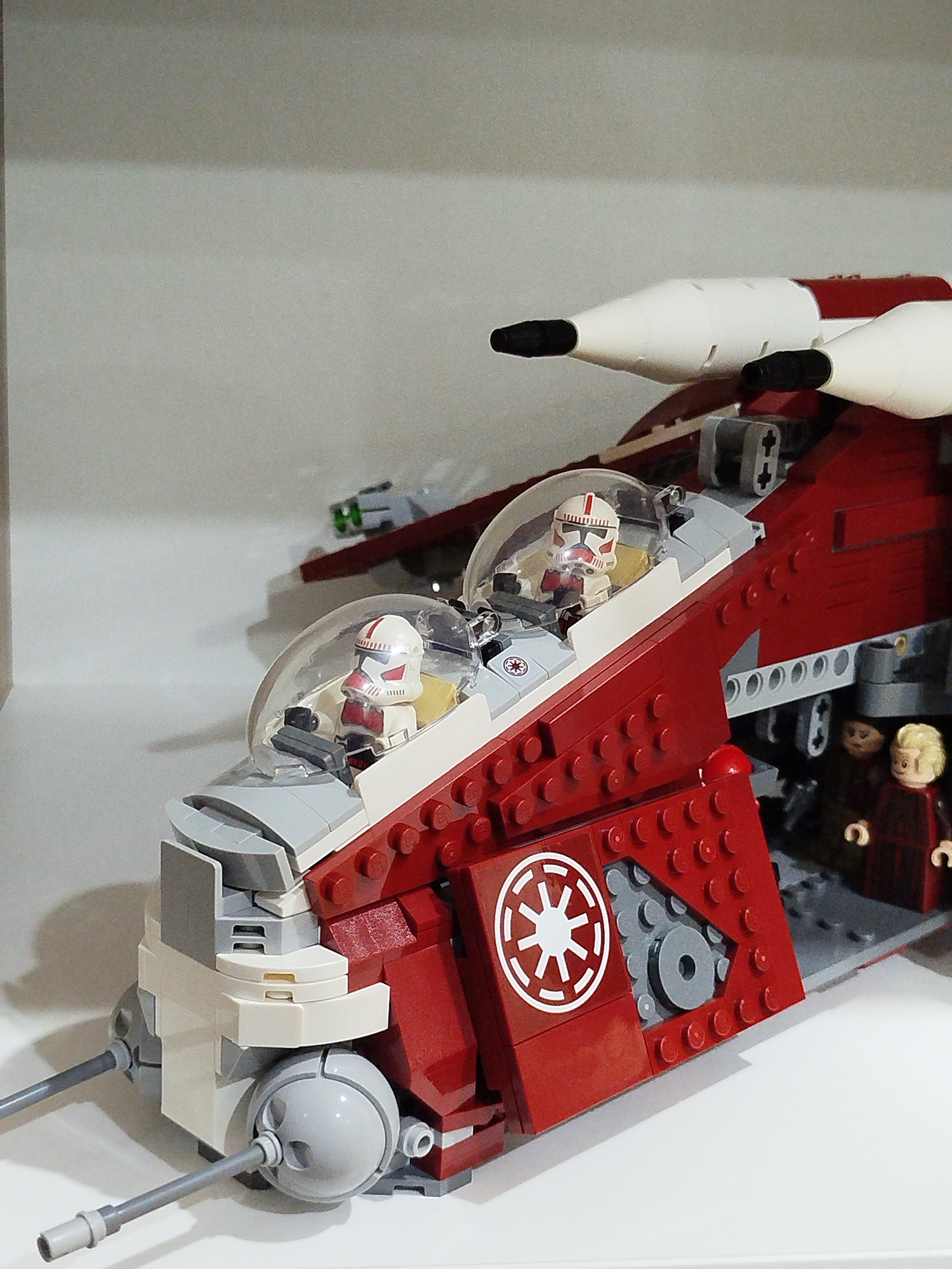 The image shows a close-up of a red and white Lego spaceship model with two Lego clone troopers in white and red helmets inside the cockpit. The spaceship has two white engine nacelles on top and is adorned with the Imperial symbol on its side. In the background, out of focus, are two Lego minifigures of what appear to be women. The spaceship is placed on a white surface, possibly a shelf, against a plain light-colored background. There is no visible indication or information within the image that suggests a location in the city of Mirny, Russia.