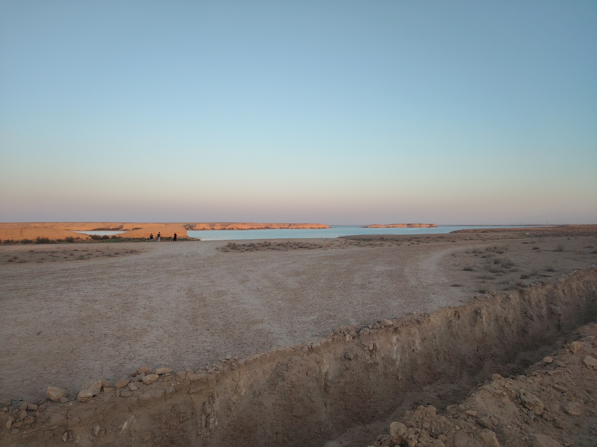 In the foreground, a trench is visible, its edges eroded and littered with rocks and soil. The ground surrounding the trench is dry and cracked, with sparse vegetation. In the mid-ground, a group of three individuals are standing on a sandy plain. Beyond them, a body of calm water, possibly a lake or sea, stretches towards the horizon. The distant shore is characterized by low, arid cliffs. The sky above is clear and exhibits a gradient of soft blues and pale pinks, indicative of either sunrise or sunset. The overall environment appears to be a desert landscape situated in or near Haratan, Uzbekistan.
