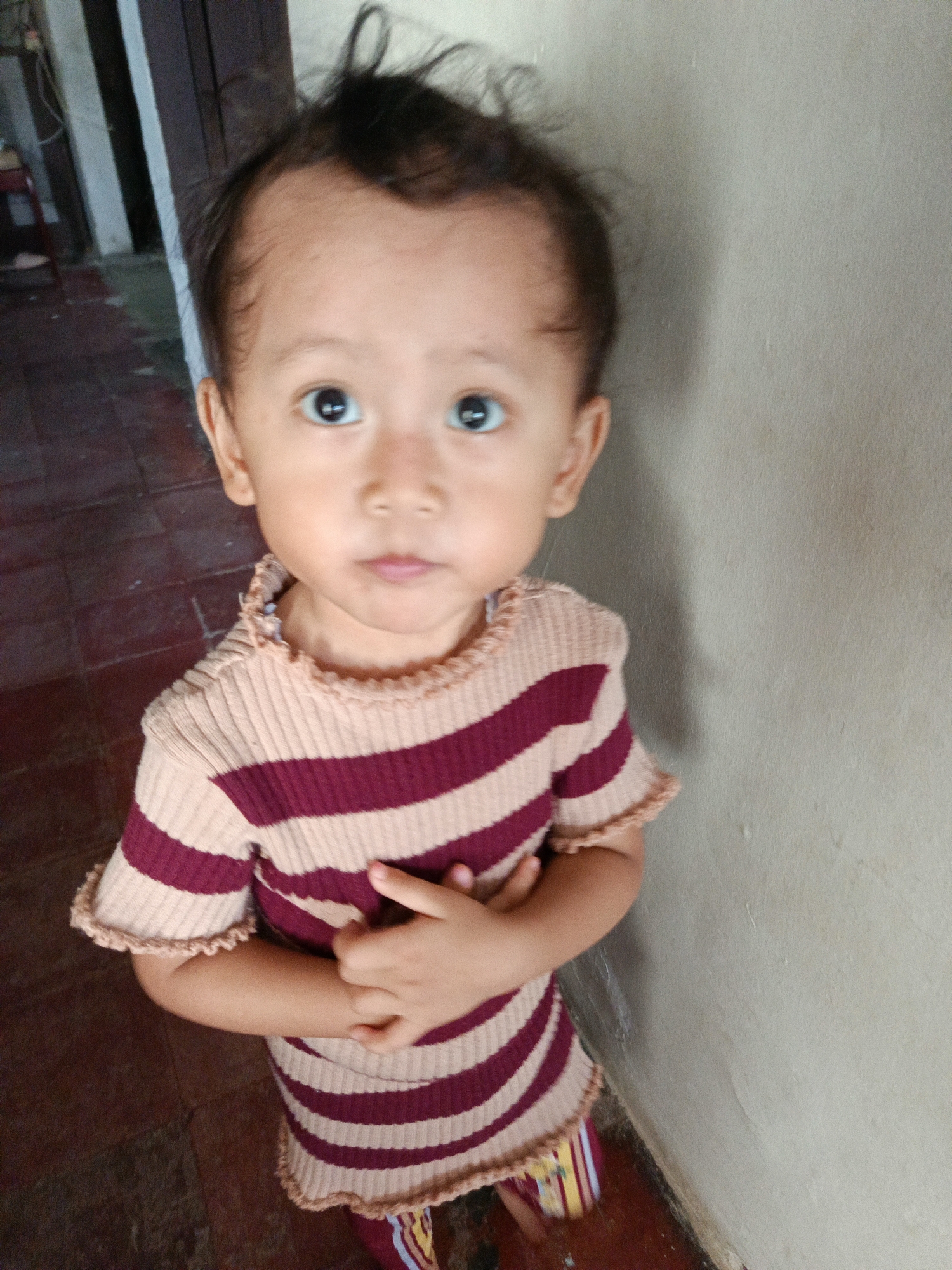 A young child stands facing the camera, their body angled slightly to the right. The child is wearing a striped shirt with alternating bands of light beige and dark pink. Their arms are crossed in front of them. The child's gaze is directed forward.
The background consists of two distinct textures. To the left, a floor made of reddish-brown tiles is visible, with a section of a dark wooden structure and some vertical white beams. To the right, a plain, light-colored wall with a slightly textured surface dominates the scene. The lighting suggests an indoor environment, possibly a home.
The overall scene appears to be an informal portrait of a child. The location in Margajaya, Indonesia, is not visually apparent from the provided image.