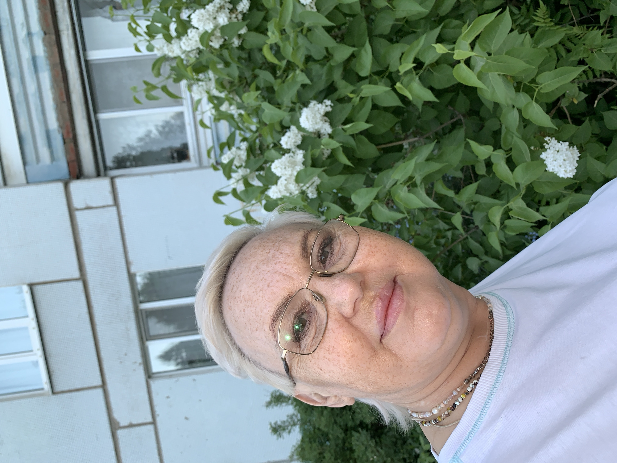 A woman with blonde hair and glasses is centered in the frame, wearing a white T-shirt and multiple necklaces. Behind her on the left is the corner of a light grey building with white-framed windows, possibly an apartment building. To the right of the woman is a large shrub with green leaves and clusters of white flowers. Based on the request, the location is presumed to be Chaikovsky, Russia.