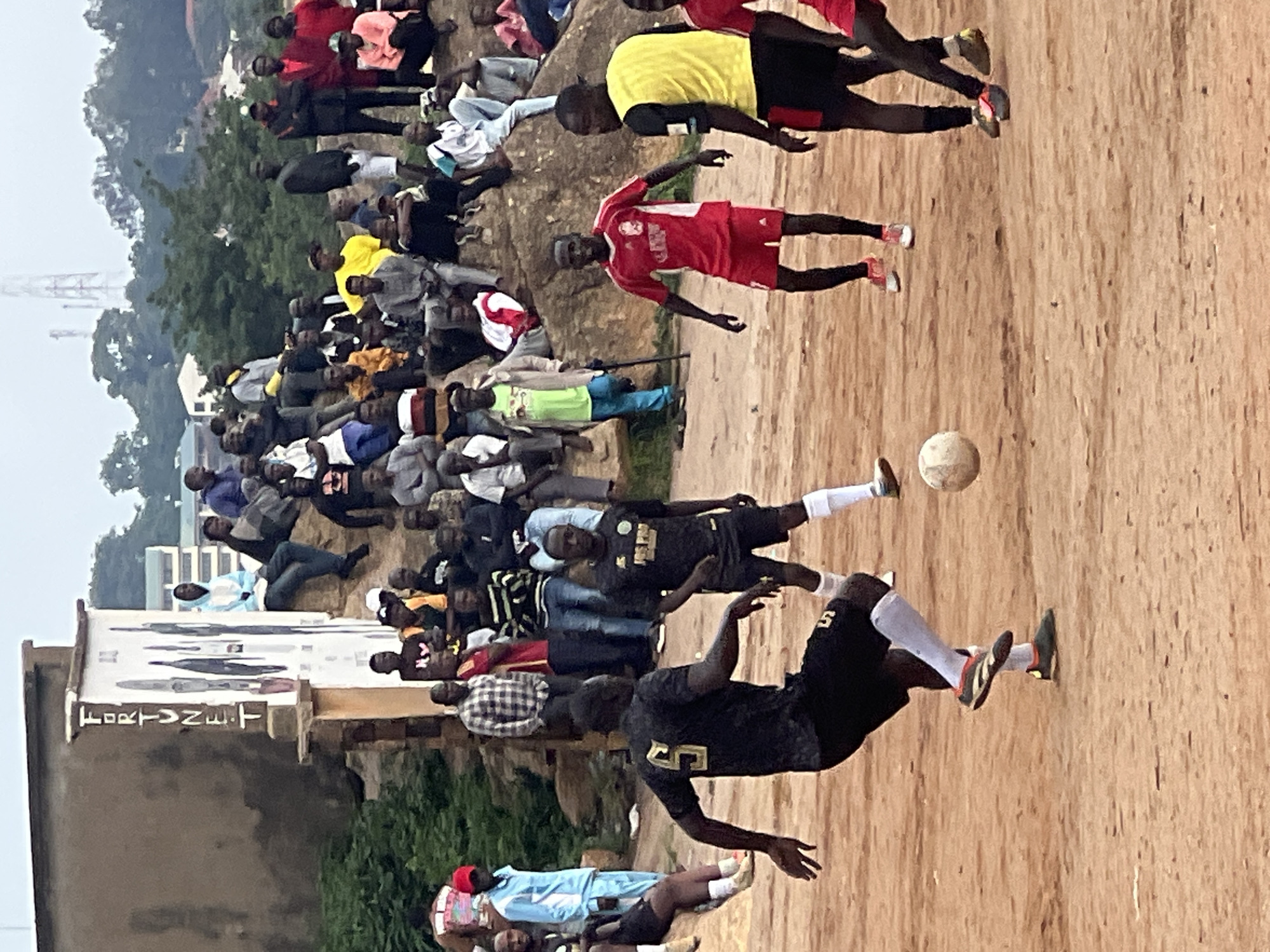 This media file captures a lively scene of a soccer game being played on a dirt field in Jos, Nigeria. A group of young men are actively engaged in the sport, with one player in mid-air attempting to kick a soccer ball. They are dressed in various soccer jerseys and shorts, with some wearing athletic socks and cleats. Spectators are gathered around the field, some sitting on the ground and others standing, observing the game. The crowd appears to be a mix of men and women, with diverse attire, suggesting a community event.
The background reveals a landscape with trees and buildings, hinting at an urban or semi-urban environment. The lighting suggests it is daytime, likely afternoon, with overcast or hazy conditions. The atmosphere seems energetic and engaged, typical of a local soccer match.
On a building in the foreground, a sign is partially visible with text that appears to read "FORTVNET". This could indicate a business or organization associated with the location. The overall impression is of a vibrant community enjoying a sporting event in an outdoor setting.