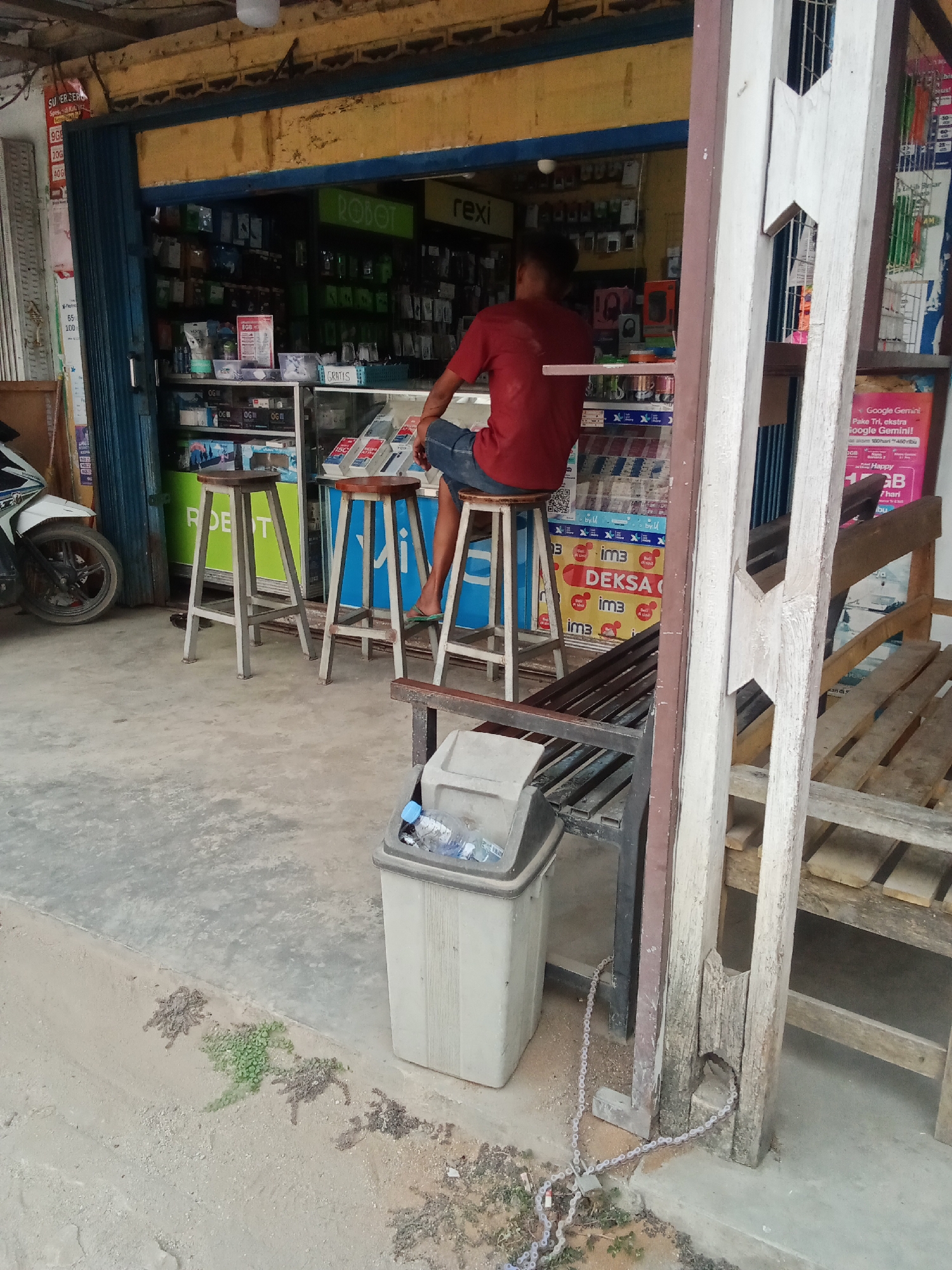 In Toto Katon, Indonesia, a man wearing a red t-shirt and blue shorts sits on a high stool in front of a storefront. The storefront appears to be a small shop selling mobile phones and accessories, with shelves stocked with various items visible through a glass display case. A motorcycle is parked to the left of the entrance, and a trash can sits to the right, containing a plastic bottle. Wooden benches are positioned near the entrance. The ground in front of the shop is paved with concrete.
