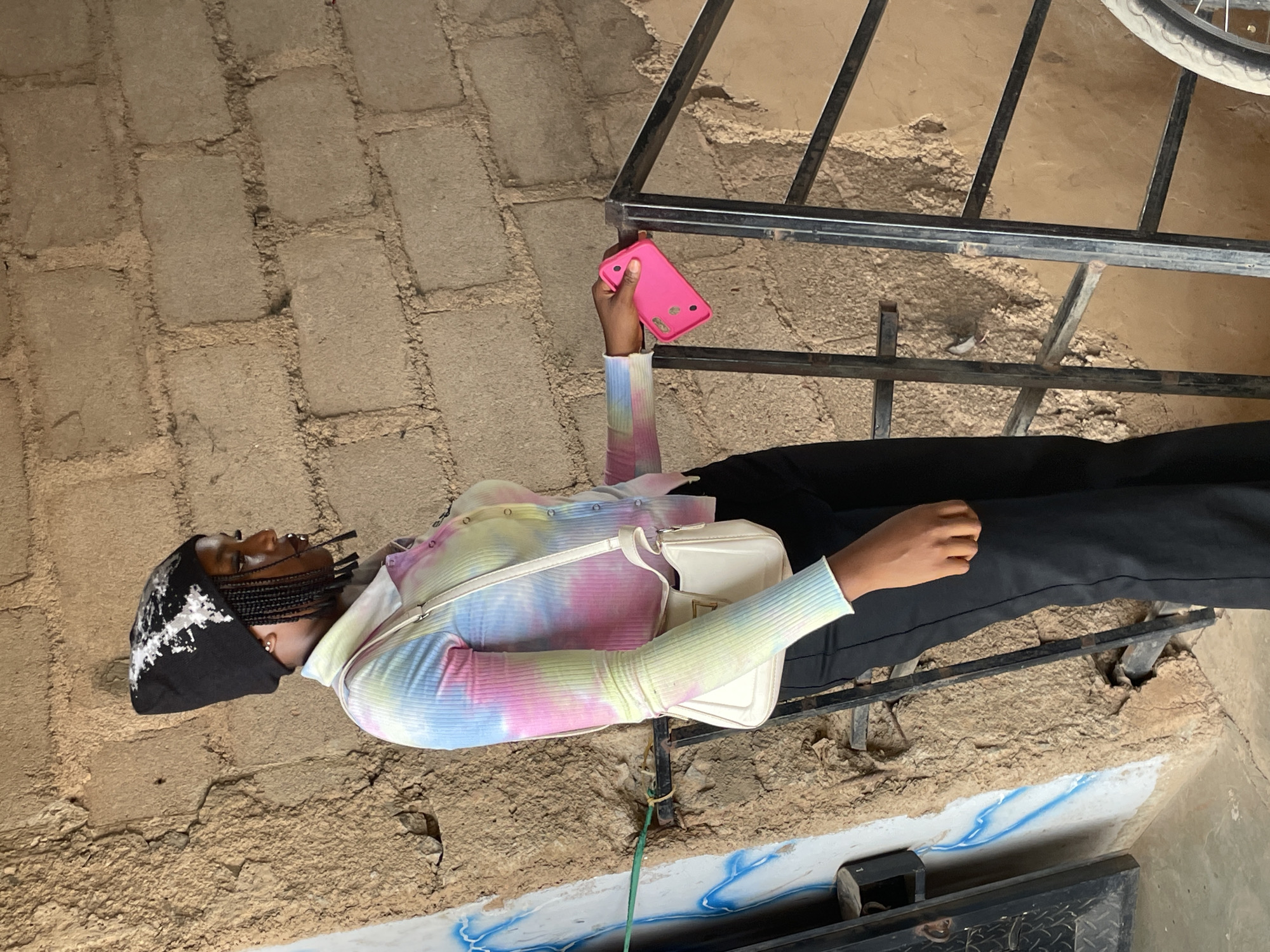 A young woman with braided hair, wearing a black beanie with white detailing, a long-sleeved tie-dye shirt, and black pants, stands with a white purse slung across her body. She holds a bright pink cellphone in her right hand. She is positioned in front of a rough, tan brick wall and a metal railing. The scene appears to be outdoors or in a sheltered outdoor area.
