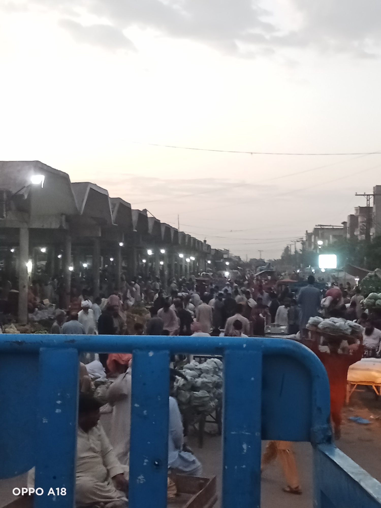 This image captures a wide-angle view of a bustling outdoor market, likely in a South Asian setting. A large crowd of people, predominantly men, fills the market street, engaged in various activities related to commerce. Many individuals are dressed in traditional shalwar kameez.
The scene appears to be set either in the very early morning or late evening, as the sky is a soft, pale gray and artificial lights within the market structures are illuminated, creating bright spots throughout the scene. The weather seems overcast.
On the left side, a long, linear structure of market stalls or buildings with distinctive, angular roofs extends into the distance, housing numerous vendors and shoppers. Each section of these structures is lit by its own overhead light. On the right side, the street is lined with more buildings and market activity, including what appears to be goods being sold from makeshift stalls.
People are seen walking, standing, congregating, and handling goods. Large quantities of produce, resembling bagged cabbage or lettuce, are visible in various parts of the market, some in piles on the ground and others stacked. One person on the far right is seen carrying a load of goods on their head. The foreground is partially obscured by a blue metal barrier, possibly the railing of a vehicle or a large cart, from behind which other people are visible.
Power lines are faintly visible crisscrossing the sky in the middle distance. The overall impression is one of a vibrant and busy marketplace. The text "OPPO A18" is visible as a watermark in the bottom-left corner, indicating the camera used to capture the image.