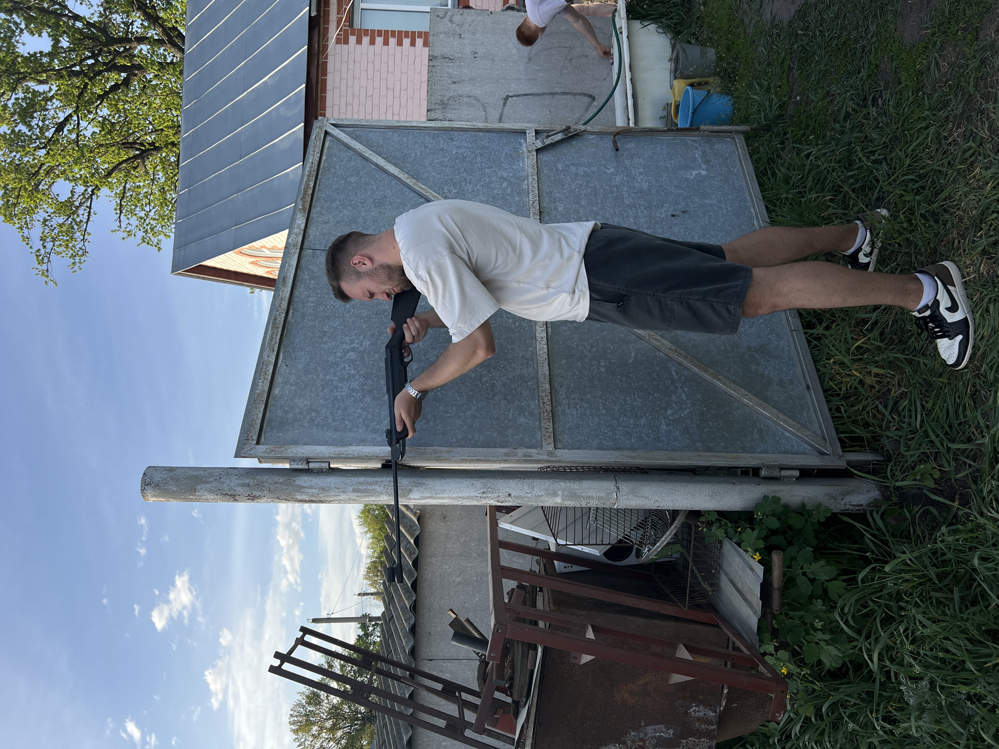 The photograph depicts a man holding a rifle outdoors. He wears a white t-shirt, dark shorts, and black and white shoes. The man is positioned in front of a large metal gate, with a small brick building visible in the background. The surrounding environment includes grass, foliage, and miscellaneous metal structures. The location is Слобода, Russia.