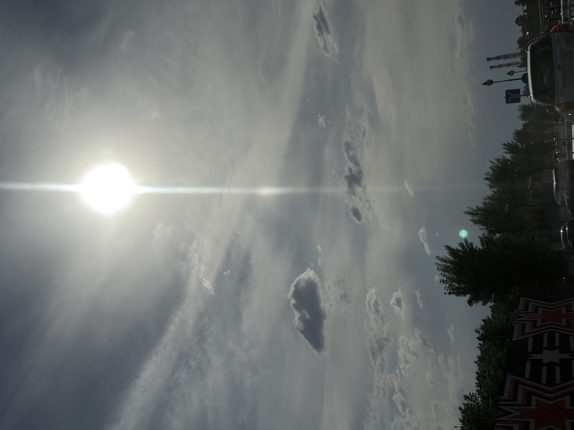 The image shows a daytime sky with thin, scattered clouds and the sun. Below, a street scene includes trees, streetlights, cars in traffic, and a building with a patterned facade. It is likely Izhevsk, Russia.
