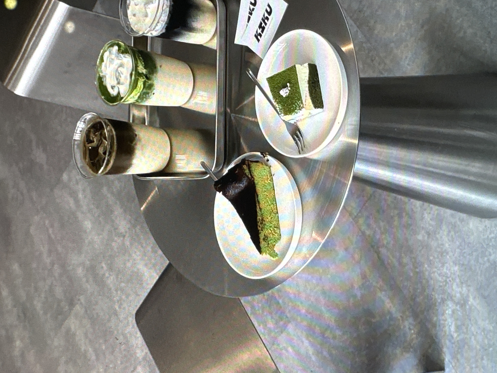 This is a close-up shot of a multi-tiered metal serving stand, likely inside a café or restaurant. On the stand, there are several beverage glasses containing layered drinks, along with two small plates. One plate holds a piece of green matcha cake with a fork, while the other plate has a wedge of green cake with a dark topping. A small paper card with the word "K3KU" printed on it is positioned near the upper plate. The stand itself is futuristic in design. The environment appears to be clean and modern. The location is Yerevan, Armenia.