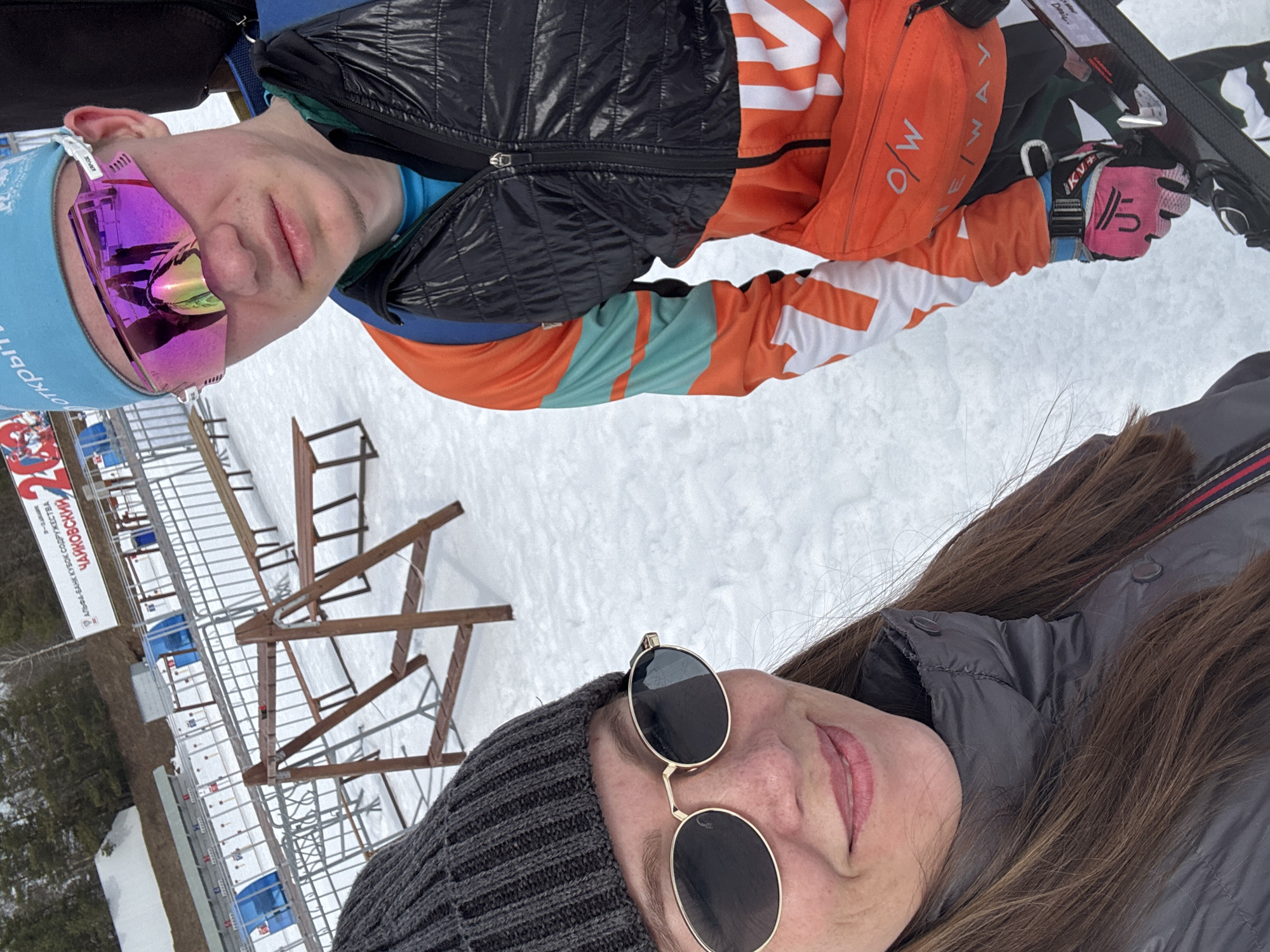 The scene depicts a selfie taken at a snowy outdoor biathlon or cross-country skiing facility, identified by a banner as hosting "Альфа-Банк Кубок Содружества Чайковский" (Alfa-Bank Commonwealth Cup Tchaikovsky). This event is located in the city of Прикамский, Russia. The ground is covered in white snow under an overcast sky.
In the lower left foreground, a female subject, estimated to be in her mid-30s to 40s, is taking the photograph. She has long brown hair, wears a dark gray knit beanie, dark oval sunglasses with gold frames, and a dark gray quilted jacket with a high collar. A subtle smile is visible on her lips.
To her right, slightly behind and occupying the right side of the frame, stands a younger male subject, appearing to be in his late teens or early twenties. He wears a light blue beanie, large pink mirrored sports sunglasses, and an athletic ski suit with sections of orange, black, and teal, featuring white "O/W ONE WAY" lettering. A black quilted vest is worn over the suit, and a blue backpack strap is visible over his left shoulder. An orange fanny pack with "O/W ONE WAY" branding is secured around his waist. His right hand, encased in a pink glove with "IKVL" and "U" logos, grasps a ski pole or ski equipment, with skis visible in the lower right corner.
The background reveals a winter sports complex with multiple lanes defined by metal barriers, small blue structures, and wooden benches. A large white banner with red Cyrillic text, partially obscured, displays "АЛЬФА-БАНК КУБОК СОДРУЖЕСТВА ЧАЙКОВСКИЙ" and "открытый" (Open). A rustic wooden A-frame structure is positioned in the mid-background. Beyond the facility, a forested area with pine and bare deciduous trees extends up a gentle slope.