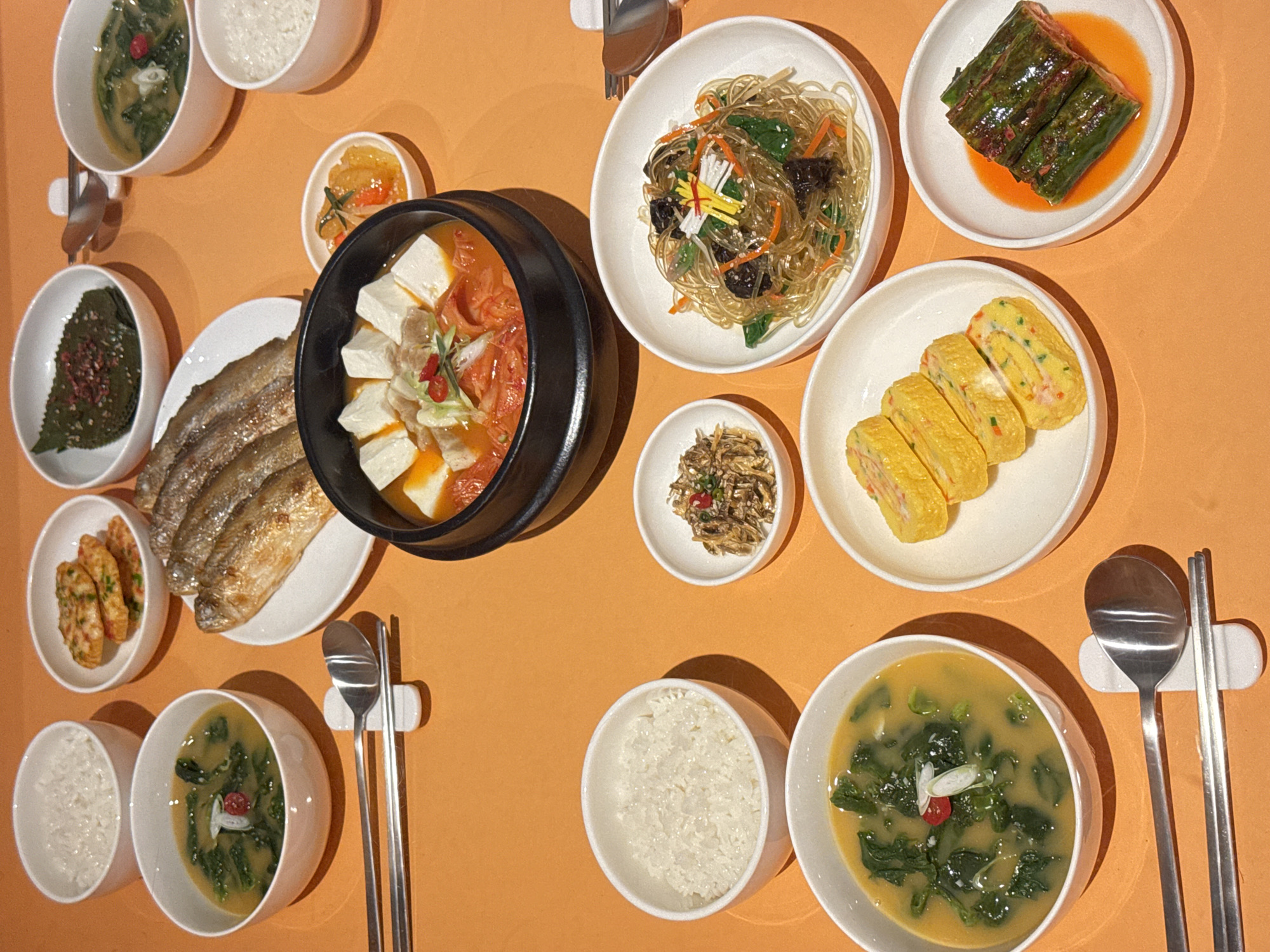 This image captures a top-down view of a Korean meal laid out on an orange surface. The spread includes several dishes, suggesting a communal dining experience. In the center, a large black pot contains a stew with tofu and kimchi. To its left, a plate holds three grilled fish. Various side dishes, or banchan, are arranged around the main courses. These include a green vegetable dish with red flakes, a small bowl of marinated dried anchovies, rolled omelets sliced into rounds, and a pickled cucumber dish. Two bowls of white rice and two bowls of soup, one a green leafy broth and the other a lighter colored broth, are also visible. A set of chopsticks and a spoon rest on a white ceramic holder. The overall presentation suggests a home-cooked meal or a casual restaurant setting in Hanoi, Vietnam. The lighting appears to be artificial, possibly from overhead, casting soft shadows on the orange surface, implying it could be daytime or evening. There is no visible text in the image.