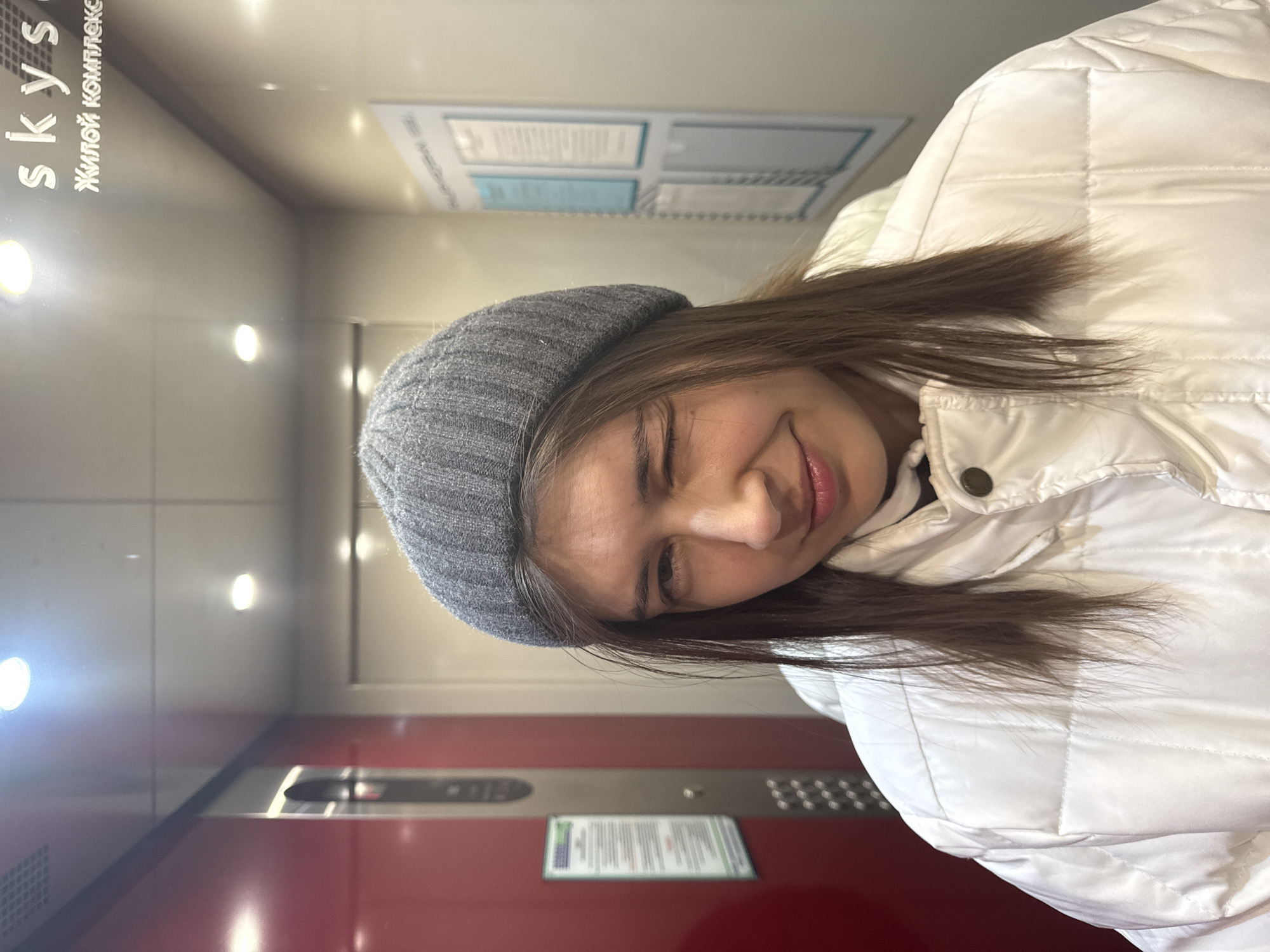 A person wearing a grey knit cap and white puffer jacket is in an elevator. The elevator's wall is red near the bottom, with a control panel featuring buttons and a digital display. A sign is positioned above the control panel. On the left side of the elevator, text in a vertical orientation reads "skys" and "Жилой Комплекс" in Russian, likely indicating the name of the residential complex. Another sign with written information is visible on the back wall of the elevator. Based on the information available, the location is likely in a residential complex called "Skys" in the city of Krasnoyarsk, Russia.