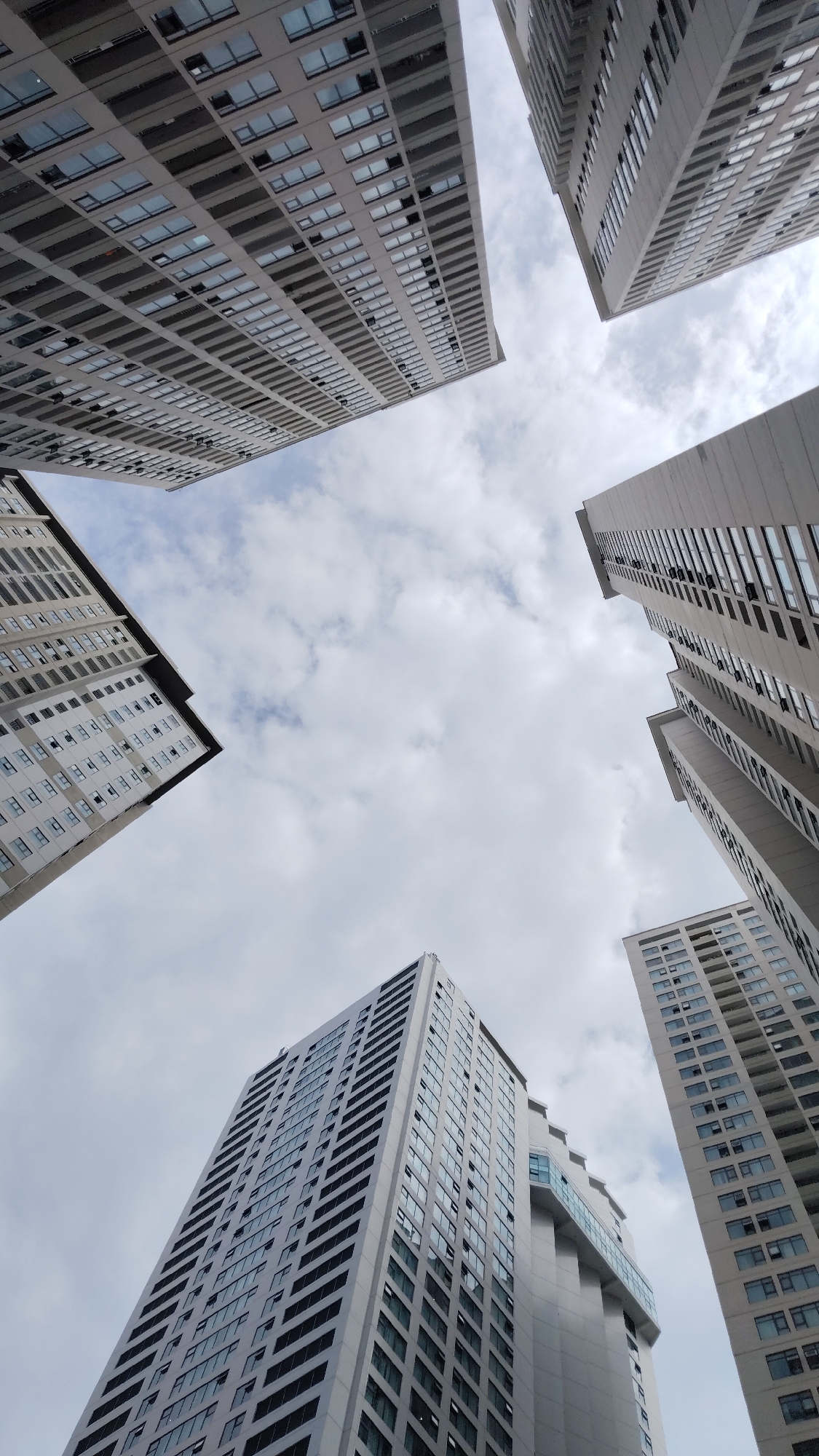 The image shows a low-angle view of several tall buildings in Bac Nha Trang Ward, Vietnam. The buildings are primarily gray or off-white, and appear to be residential apartment buildings. The sky is cloudy and visible in the spaces between the buildings. There are no people visible in the image.