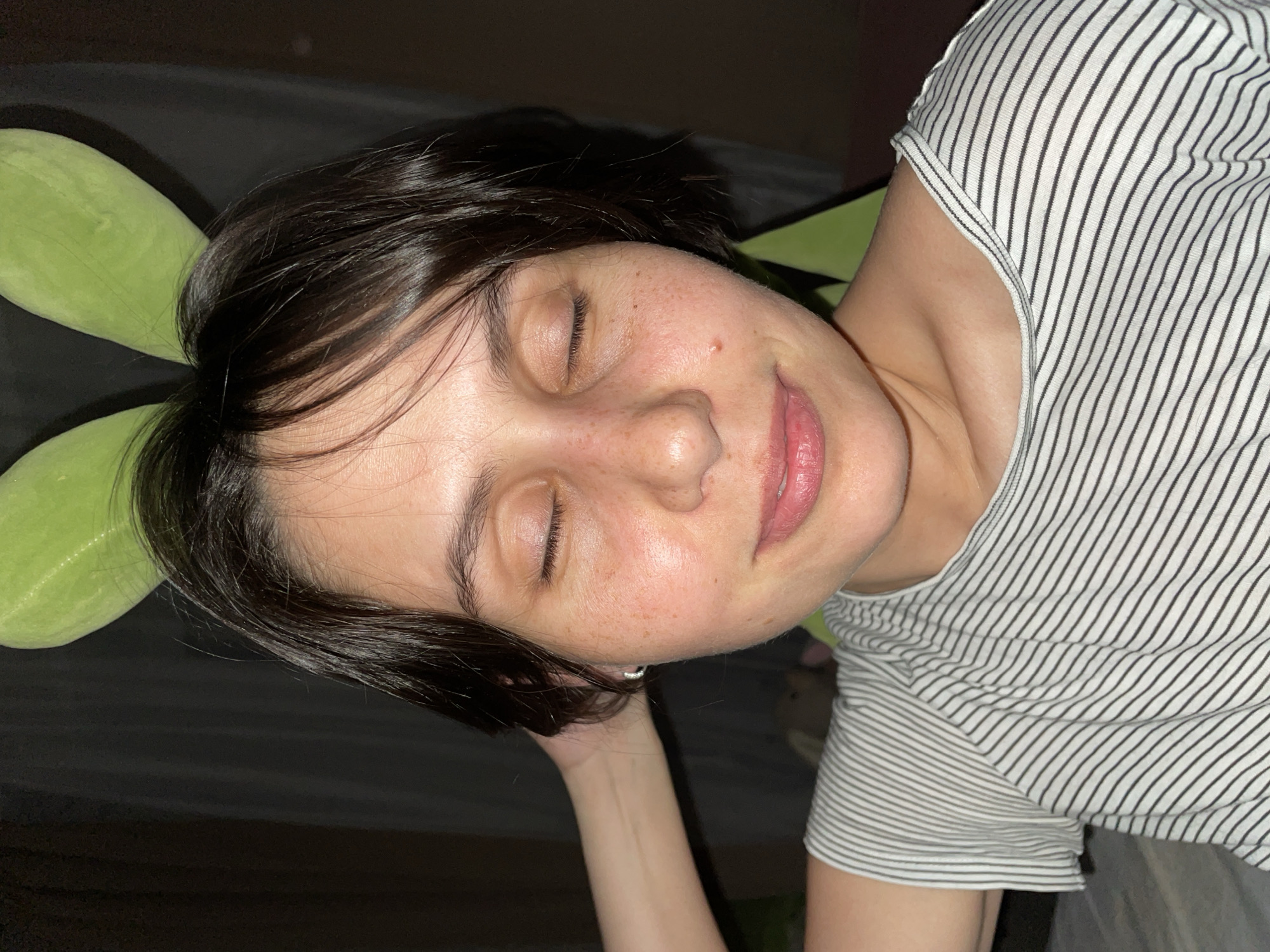 A woman with short, dark hair and freckles is lying down with her eyes closed, appearing to be sleeping or resting. She has fair skin and is wearing a striped shirt. Next to her head is a green, elongated object resembling a pillow or stuffed animal. The background appears to be a dark, possibly indoor environment. There is no information in the provided image to indicate this scene is located in Izhevsk, Russia.