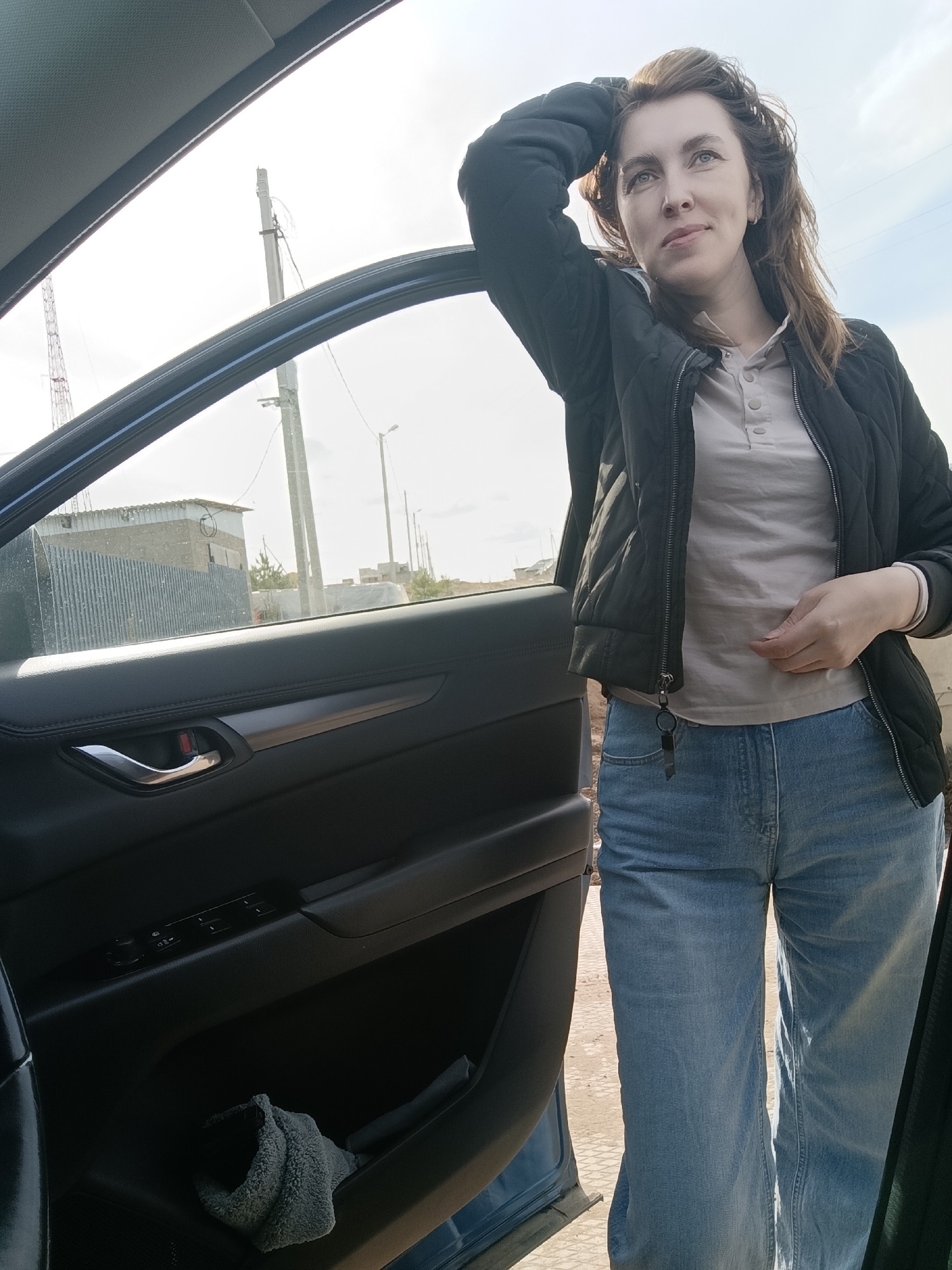 A woman stands next to an open car door. She is wearing a black quilted jacket, a light gray button-down shirt, and blue jeans. Her right hand rests on her stomach, and her left arm is bent with her hand touching her head. The car door is black with silver trim and a cloth in the pocket. Outside the car, there are buildings, utility poles, and the sky. Based on visual markers, this image was taken in the city of Chaikovsky, Russia.