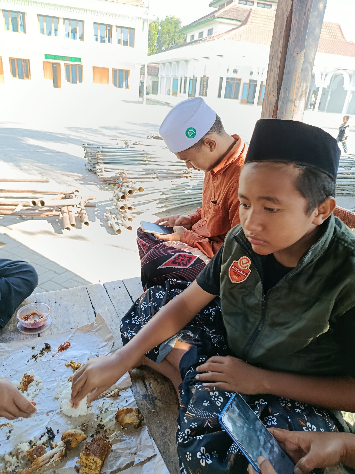 In Ramban Wetan, Indonesia, two young males are seated on a wooden platform, engaged in eating and using mobile phones. The male on the left, wearing an orange shirt and a white peci (cap), is looking down at his phone. The male on the right, wearing a dark green vest over a black shirt and a black peci, is also looking at his phone and reaching for food on a piece of brown paper spread on the wooden surface. Scattered on the paper are portions of white rice, fried chicken pieces, and a small dish containing a red condiment. Several other individuals, whose faces are not clearly visible, are also present and interacting with the food. In the background, a white building with arched windows and a red-tiled roof, likely a mosque or educational institution, is visible. Piles of metal scaffolding are stacked on the ground in the middle ground. The scene is outdoors, under a bright sky, casting shadows on the ground.