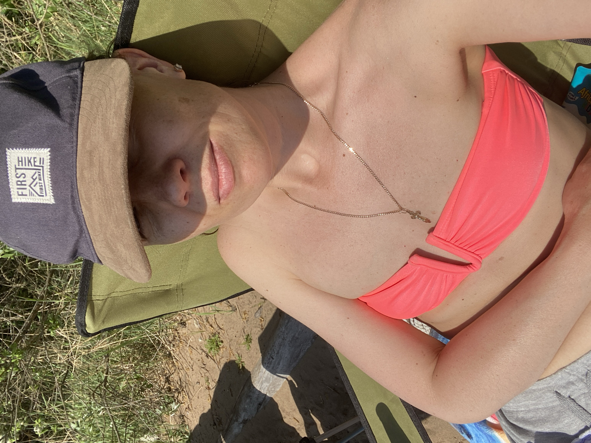 A woman lies in a green camping chair in an outdoor setting. She is wearing a pink bikini top and a grey baseball cap with a patch that says "HIKE FIRST HIKE." A thin gold chain with a cross pendant is around her neck. Grass and shadows are visible in the background, and the chair appears to be on a patch of dirt or sand. There is no information in the image to identify the location as the city of Chaykovsky, Russia.