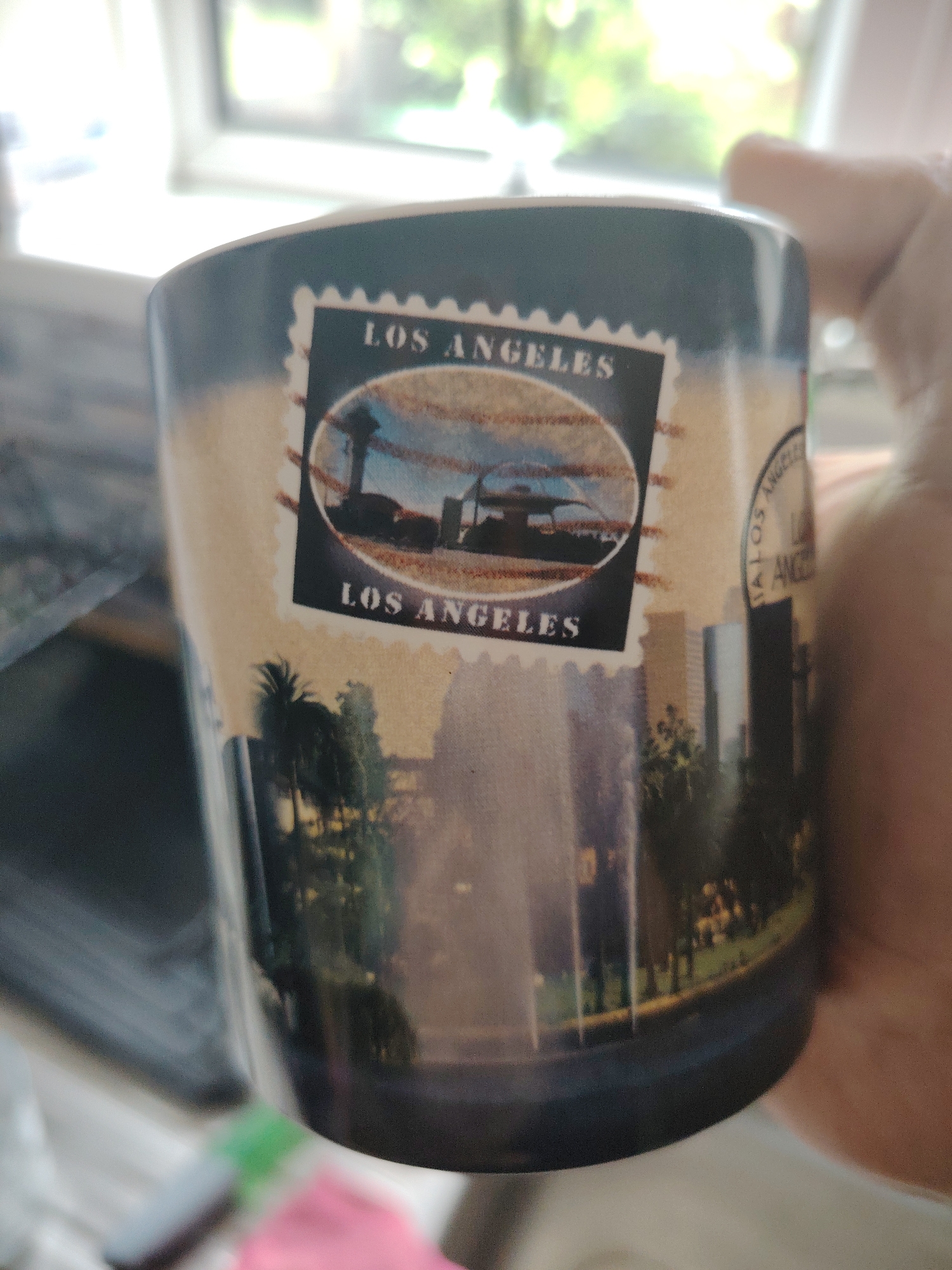 A close-up, eye-level shot captures a ceramic mug held vertically by a person's light-skinned hand on the right side. The mug features a prominent stamp-like design with "LOS ANGELES" printed at the top and bottom. Inside this stamp is an oval image depicting the Griffith Observatory or a similar structure, alongside palm trees against a cloudy sky. Below this, a panoramic cityscape wraps around the mug, showcasing tall buildings and palm trees with a fountain or water feature in the foreground. A partial circular seal with text, likely "LOS ANGELES," is visible on the right edge of the mug. The background is brightly lit and out of focus, revealing a window with green foliage visible outside, suggesting an indoor setting. Blurry household items in shades of pink and green are faintly visible at the bottom left.
