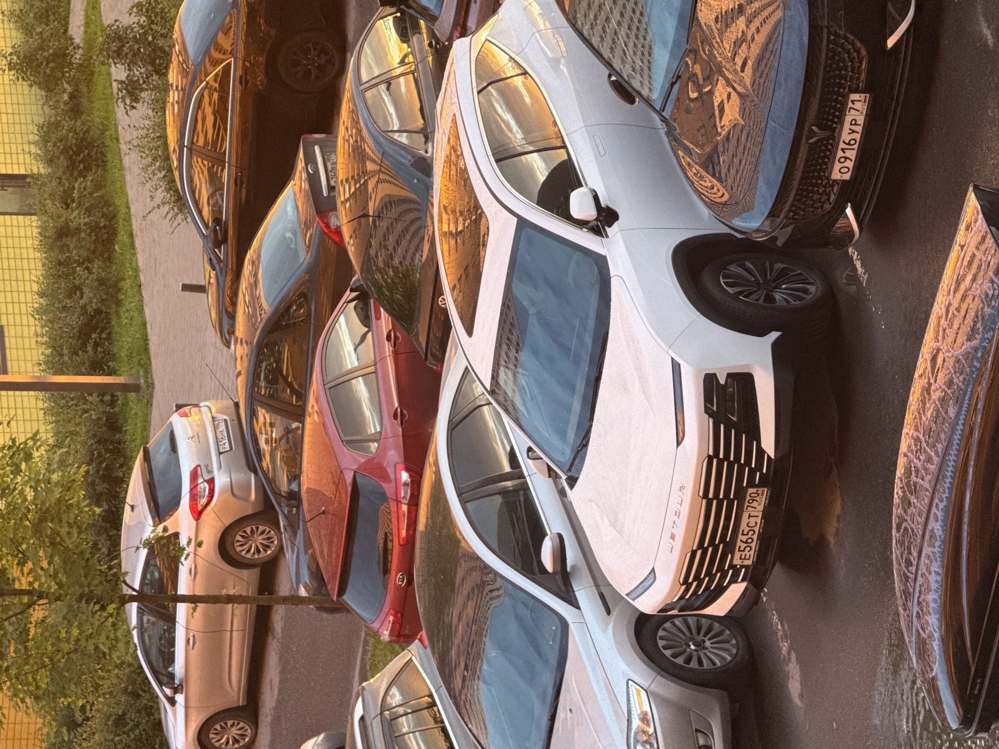 This image depicts a parking lot in Drozhzhino, Russia, bathed in the warm glow of sunset. Several cars are parked, including a silver hatchback with a license plate starting with "P416HK", a red hatchback with "KIA" badging, a dark sedan reflecting the sunset, and a white SUV with a license plate "E565CT 790". In the foreground, a dark roof box is visible. The scene is surrounded by green foliage and a building with a yellow, textured facade. The lighting suggests it is late afternoon or early evening, casting long shadows and highlighting the metallic surfaces of the vehicles.