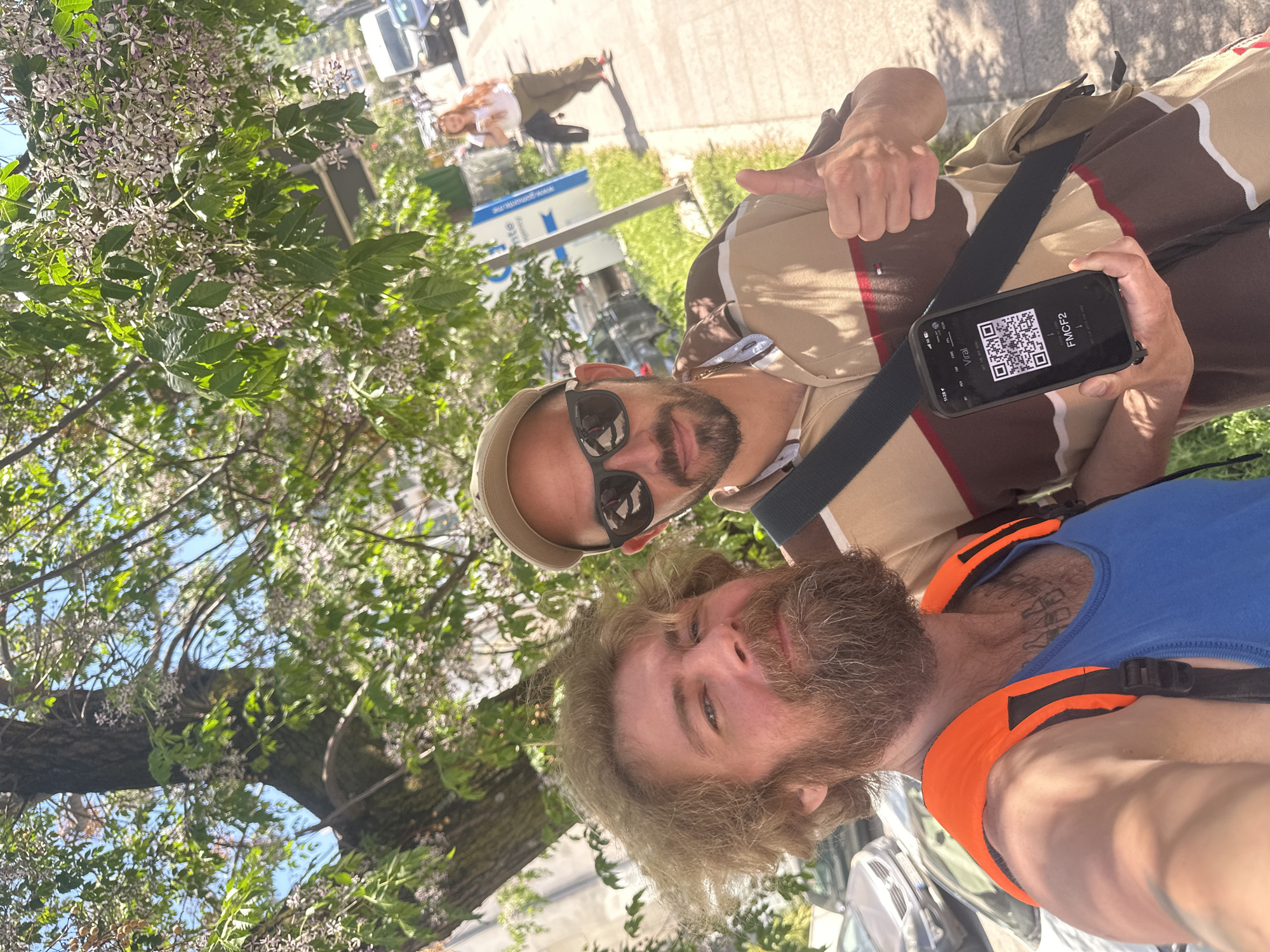 The image is a bright, sunlit selfie taken outdoors, featuring two men. The camera is rotated, giving the impression of an upward diagonal angle.
In the foreground on the right, a man with a beard and brown hair, wearing a blue tank top with prominent orange shoulder straps, looks towards the camera. To his left and slightly behind, another man is visible, wearing a beige and brown striped polo shirt, a light-colored cap, and dark sunglasses. He holds a black smartphone in his left hand, displaying a QR code on its screen. The phone screen also shows the text "VIRAL", "FMCF2", and the time "11:55". This man is simultaneously giving a thumbs-up with his right hand. Both men appear relaxed and friendly.
The background shows an outdoor setting with a large tree featuring abundant green leaves and clusters of small, pale purple flowers, suggesting a sunny day in a natural or park-like area within an urban environment. Bright sunlight casts shadows, indicating it is likely midday or early afternoon. In the further background, out of focus, are faint outlines of buildings, other people, and what appear to be vehicles. A blue sign in the distance prominently displays "www.montenegro.me", confirming the location as Budva, Montenegro.