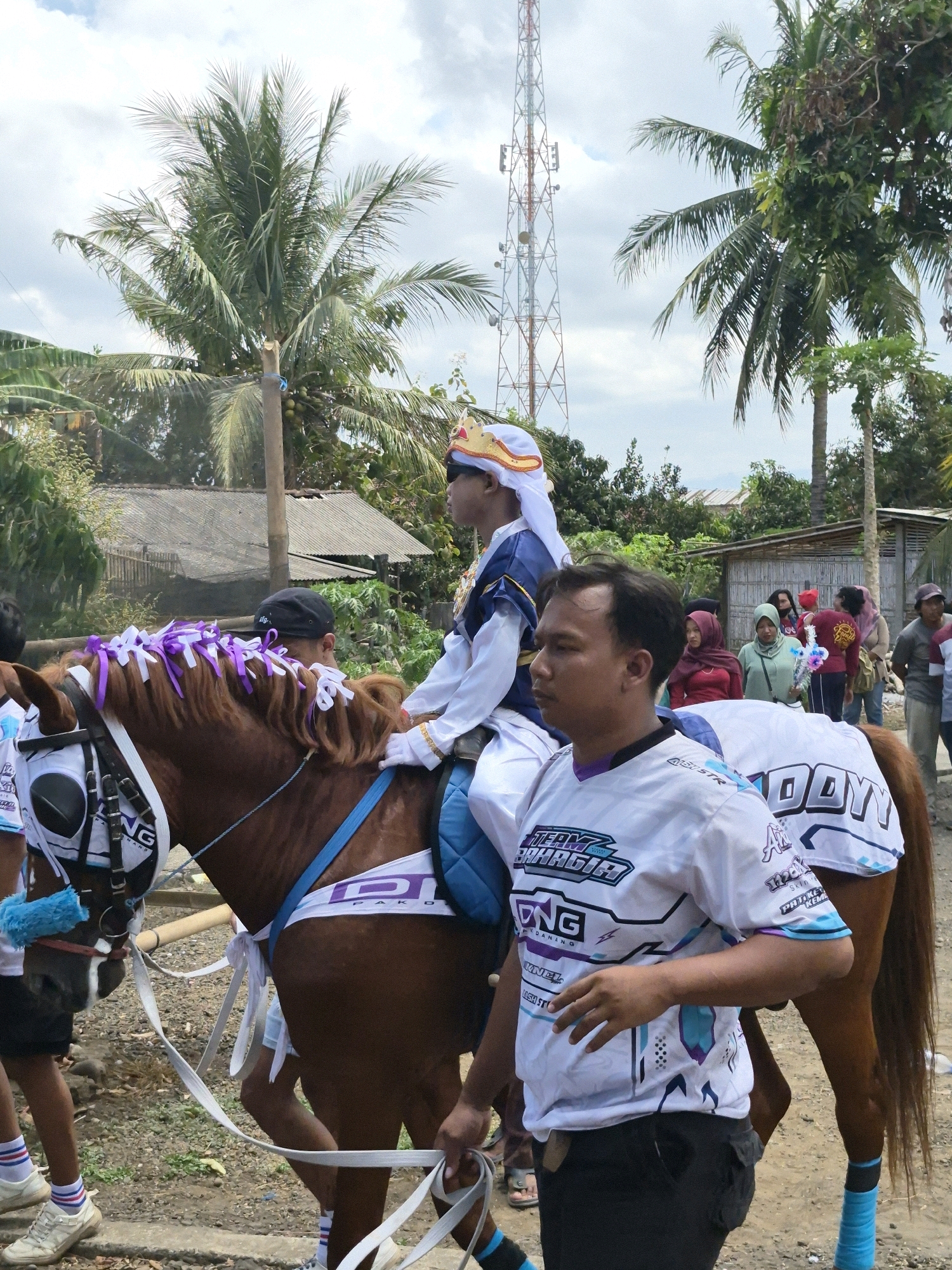 In Grantung, Indonesia, a brown horse, adorned with purple and white ribbons in its mane and blue and white decorative coverings, is being led by a man wearing a white t-shirt with a graphic design and black pants. On the horse's back sits a young person dressed in a blue and white costume with a golden crown. Behind them, a crowd of people, including women in headscarves, observe the scene. The background features a telecommunications tower, palm trees, and simple houses, indicating a rural or suburban environment. The sky is partly cloudy.