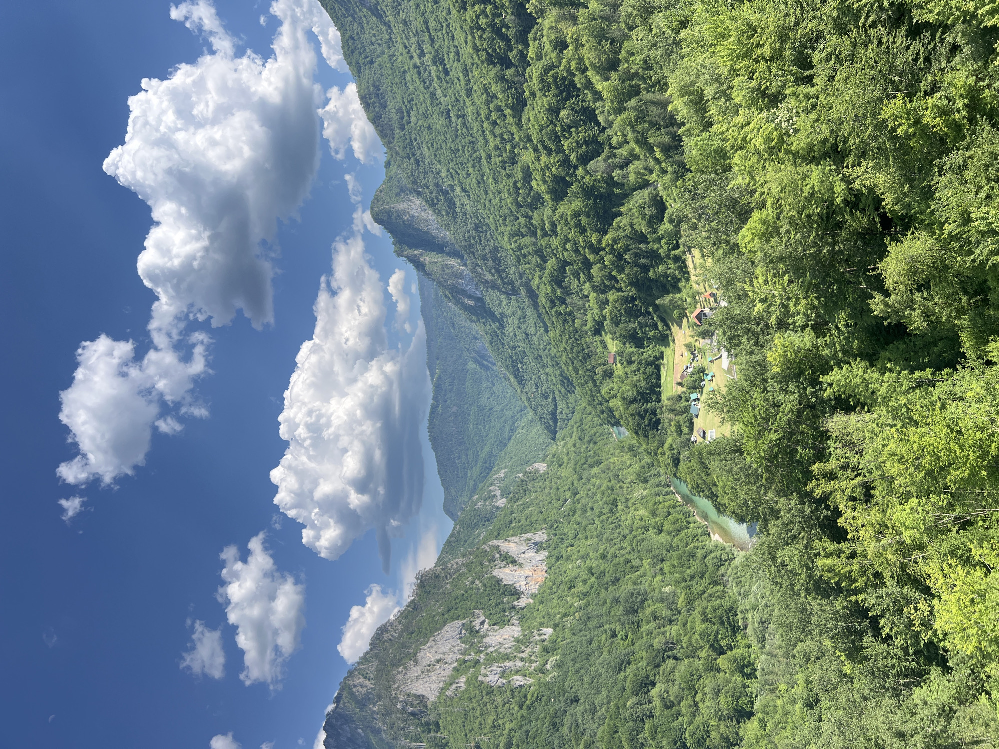 The image displays a scenic view of the Tara River Canyon in Đurđevića Tara, Montenegro. The canyon is characterized by steep, tree-covered mountains on either side, with a turquoise river flowing through the valley below. The vegetation is lush and green, indicative of a temperate climate. The sky is blue with scattered white clouds. In the mid-ground, nestled amongst the trees, are a few small buildings, suggesting a rural settlement. The overall impression is one of natural beauty and tranquility.