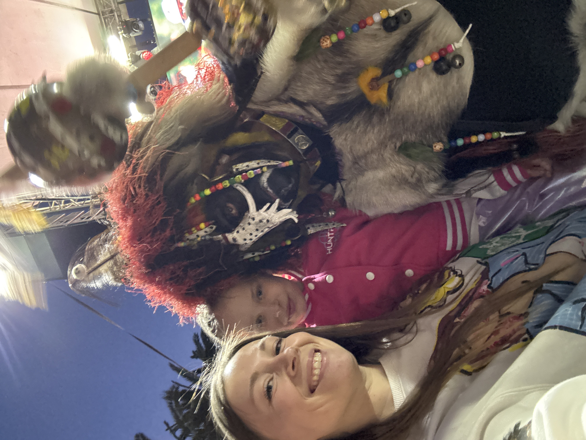 A selfie-style photograph taken from a low angle features a woman, a child, and a large, ornate mask or costume. The woman has light skin and brown hair, and she is smiling broadly at the camera. The child, also with light skin, is wearing a pink jacket with the word "HUNTER" visible. The mask/costume is elaborate, featuring red fringe, fur, and colorful beadwork. The background shows elements of a city scene at dusk or night, with streetlights and palm trees visible. Due to the angle and limited background detail, the specific location in Budva, Montenegro, cannot be determined with certainty.