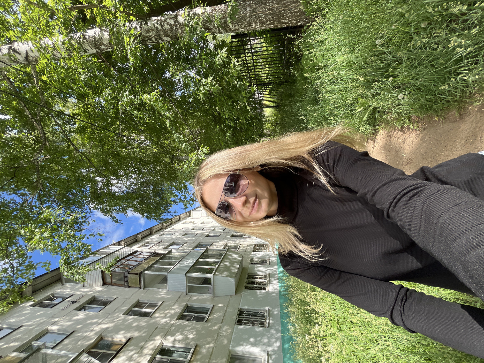 The image is a selfie captured in a portrait orientation, then rotated 90 degrees clockwise, making the horizon appear vertical. In the foreground, a blonde woman is seen from the chest up, wearing large, dark sunglasses and a black ribbed turtleneck or long-sleeved top. Her blonde hair flows to her left, indicating some movement or breeze. Her expression is largely obscured by the sunglasses.
The background is split between a residential building and a natural green environment. To the left of the woman (if the image were upright), a multi-story, light-colored building with numerous windows and enclosed balconies extends towards the upper left corner. Parts of the building's facade appear to have a light green lower section. Above the building, a clear blue sky with some white clouds is visible, framed by green tree leaves.
To the right of the woman (if the image were upright), a lush green area with trees, including a prominent birch tree with its distinctive white bark, dominates. Through the dense foliage, a dark metal fence is partially visible. Below the trees and to the right of the woman's body, a patch of green grass with small white flowers (resembling dandelions) borders a light-colored dirt path or track.
The scene is well-lit by natural sunlight, suggesting a bright daytime. Shadows from the trees are cast on the building. The overall setting is outdoors, likely a park or courtyard next to a residential area in Chaikovsky, Russia. There is no visible text in the image.