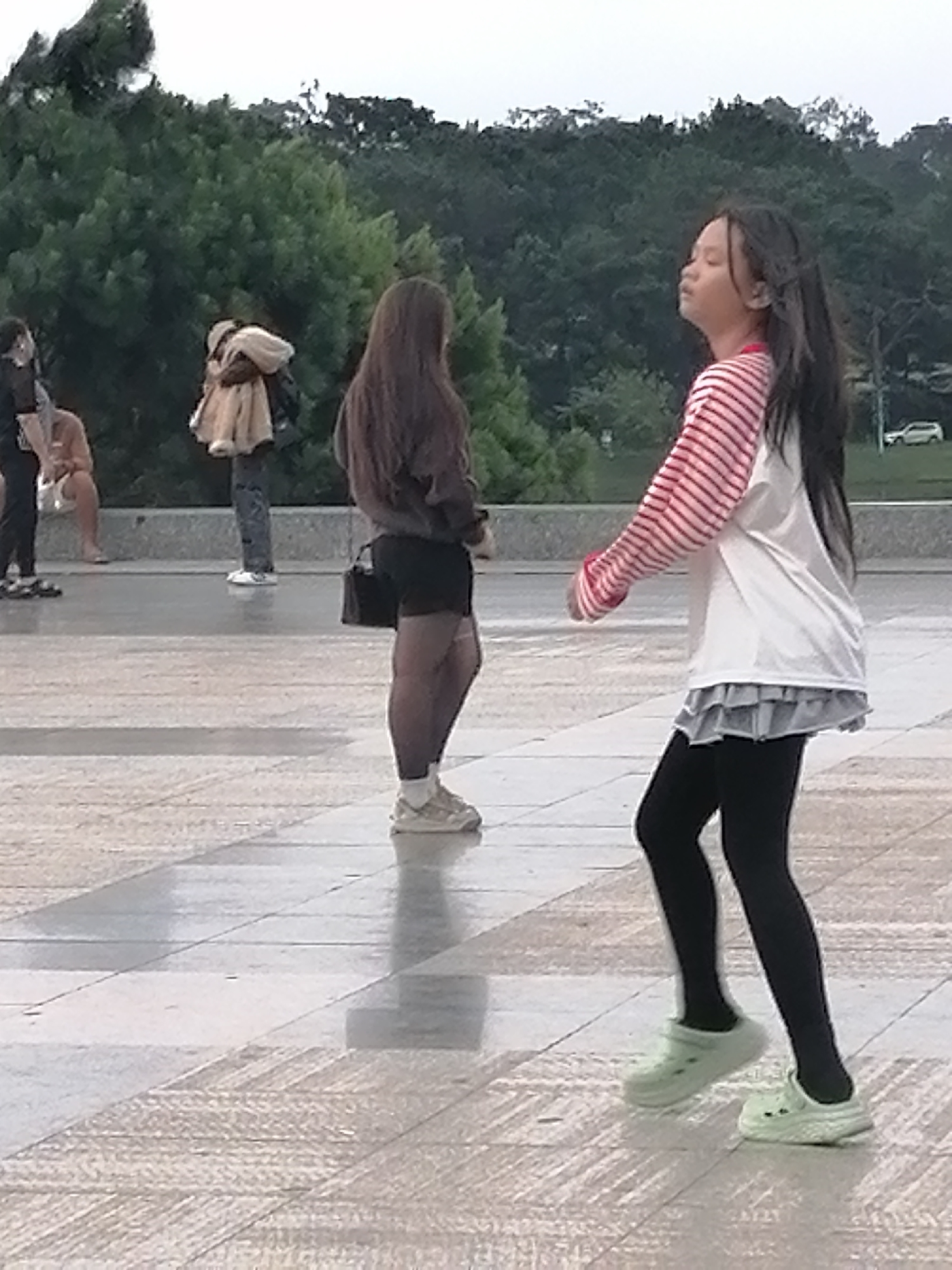 The image shows a group of people in an outdoor setting. In the foreground, a young girl with long dark hair is walking and looking upwards, wearing a white long-sleeved shirt with red stripes, a grey ruffled skirt, black leggings, and light green clogs. To the left and further back, another person is standing with their back to the camera, wearing a brown long-sleeved shirt, black shorts, patterned tights, and white sneakers. In the background, two individuals are visible: one is wearing a light-colored jacket over dark pants, and another person is seated. The ground is paved with stone tiles that appear wet. Lush green trees form the backdrop, suggesting a park or garden area. There is a white car in the far background. This scene is likely located in the city of Da Lat, Vietnam, known for its scenic parks and gardens.