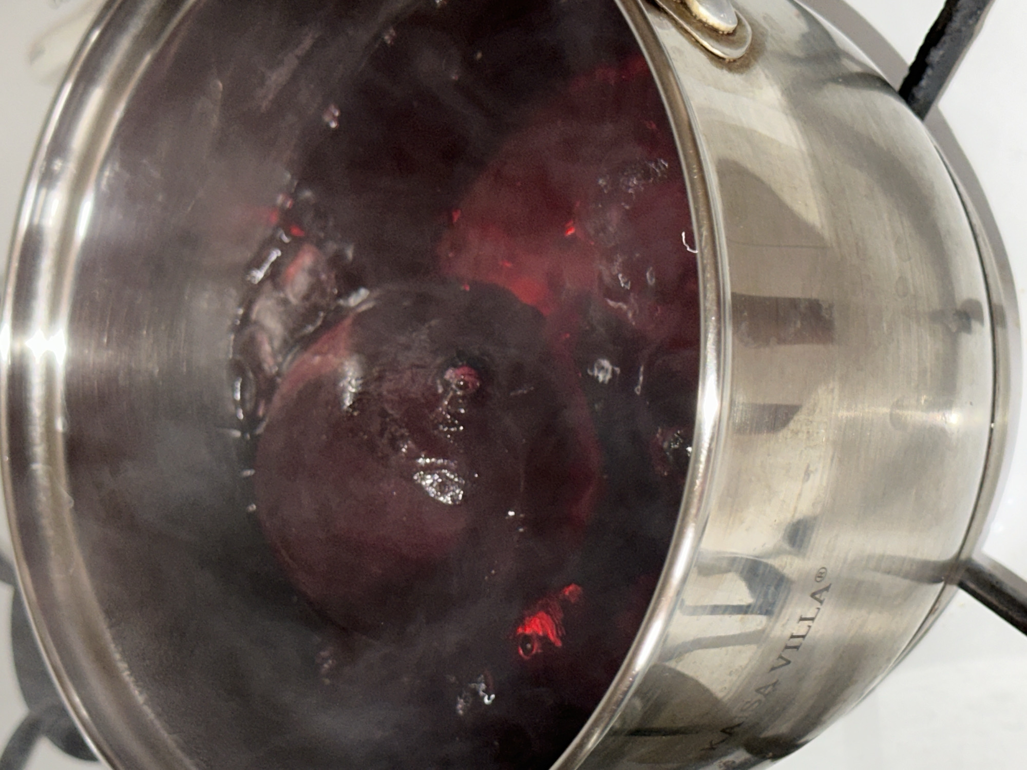 A close-up, overhead shot shows whole beets boiling in a stainless steel pot filled with dark red liquid. The pot is sitting on a white stovetop, and steam rises from the boiling contents. The pot has a textured band around its rim and the brand name "KAISA VILLA" is visible in the lower left quadrant. There are no people or other objects clearly visible in the frame.