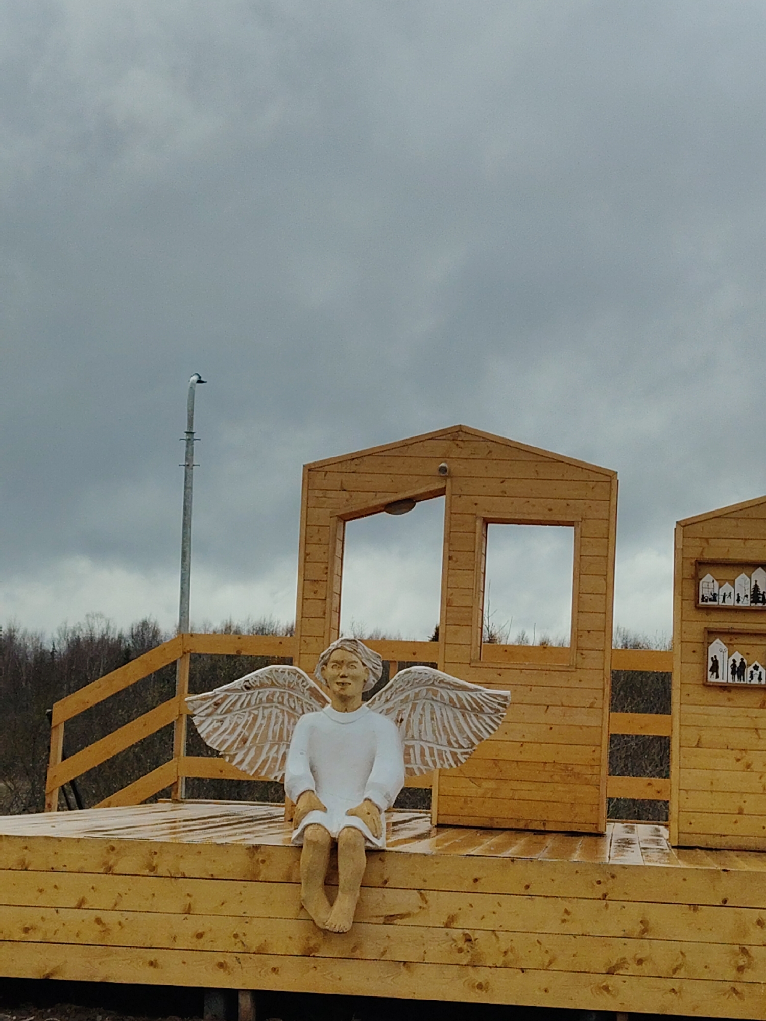A full shot depicts a wooden platform with a small building and an angel statue. The sky is overcast and gray.
The platform is constructed of light-colored wood, with a small wooden building behind the angel statue. The building has a window on each side of an open door. To the right, there is another small wooden structure with decorative cutouts of people and houses. A metal post with a light fixture stands to the left of the building. The angel statue is seated on the edge of the platform, with its legs dangling down. It has carved wings and is wearing a simple white dress.
In the background, there are trees and foliage. The overall setting suggests an outdoor art installation or public space.