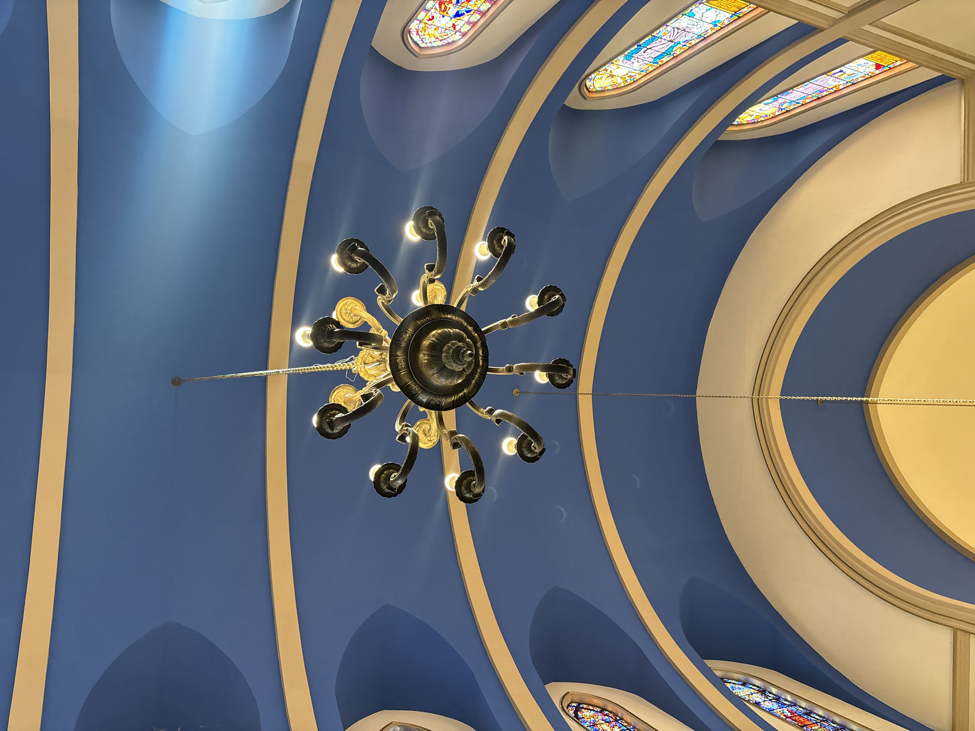The image depicts the interior ceiling of a building, likely a church or a grand hall, with a distinct blue ceiling accented by cream-colored curved trim. A large, ornate chandelier hangs from the center, featuring multiple arms with illuminated bulbs. On either side of the ceiling, tall, narrow stained-glass windows are visible, depicting colorful religious or decorative scenes. The architectural style suggests a place of worship or a historical building. The specific location in São Paulo, Brazil, is not discernible from the image alone.