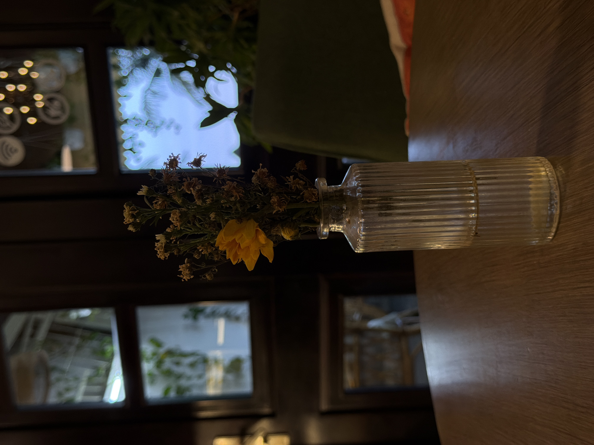 A clear, ribbed glass vase containing dried sprigs and one yellow flower lays horizontally on a wooden table. In the background, a dark wood framed window reveals an out-of-focus view of trees and the exterior of a building. Faint light fixtures are also visible through the window. The scene is lit by ambient light. The location is suggested to be Bac Nha Trang Ward, Vietnam, based on user-provided context.