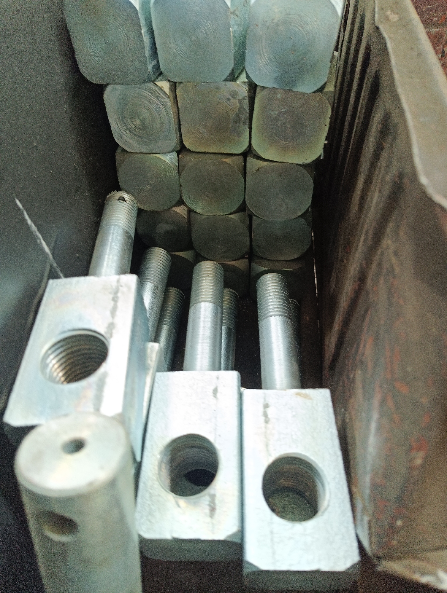 The image shows a close-up, high-angle view of various metal components stored within a dark, possibly metal, bin or box. In the background, numerous square-ended metal bars or blocks are stacked. The ends of these square pieces display concentric circular patterns, suggesting they were cut from larger stock or machined. Some of these square ends have a greenish-grey hue, while others appear darker or slightly discolored.
In the foreground, several threaded metal rods or bolts are visible. These rods are inserted into or attached to flat, rectangular metal bases, each with at least one circular hole. The bases are generally light grey, indicating a metallic finish, possibly galvanized or zinc-plated. One base in the lower left foreground has a large, internally threaded circular opening. Another base slightly to its right also has a similar large circular hole. A long, slender cylindrical metal piece with a smaller hole on its end and another on its side is partially visible in the bottom left corner. The threaded sections of the bolts are bright and metallic. The interior surface of the container on the right side appears dark and somewhat textured, possibly showing signs of rust or wear. No text is visible in the image.