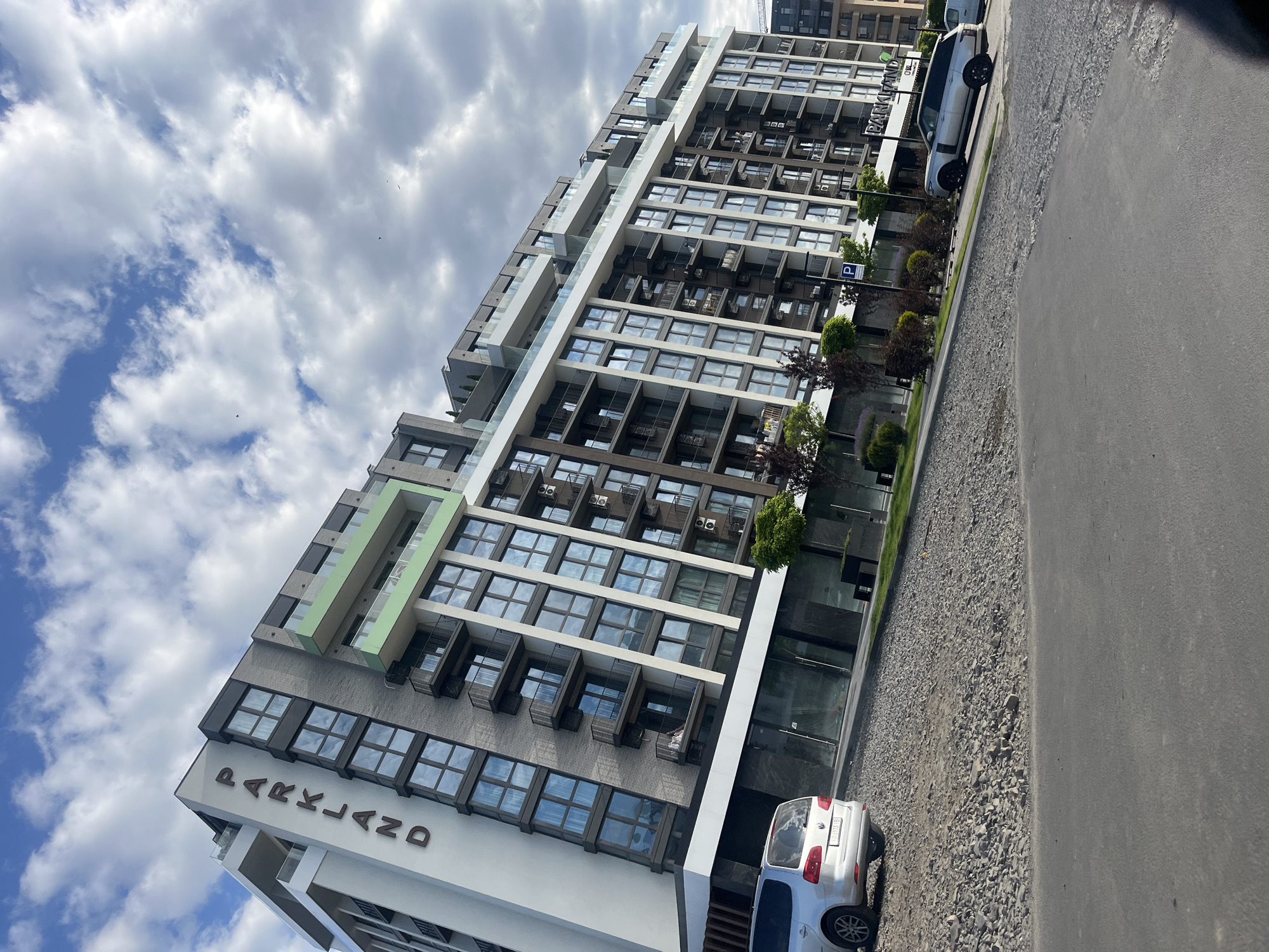The image shows a modern, multi-story apartment building with a white and grey facade, accented with light green balconies. The word "PARKLAND" is prominently displayed on the building's exterior in large, dark lettering. Several cars are parked in front of the building, including a silver Volkswagen minivan and a silver SUV. The ground in front of the building is covered with gravel and asphalt. The sky is cloudy with patches of blue. The scene appears to be in an urban or suburban setting. There are no people visible in the image.
