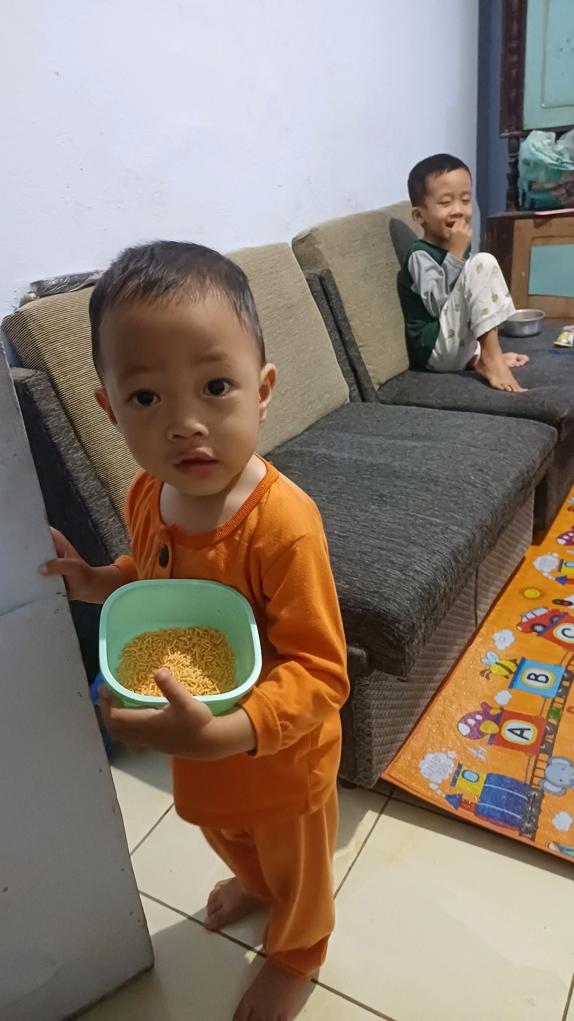A young child wearing an orange pajama set stands holding a light green bowl filled with uncooked instant noodles. The child is looking directly at the camera with a neutral expression. In the background, an older child is sitting on a sectional couch, laughing and touching their face. The older child is wearing a gray and green long-sleeved shirt and patterned pajama pants. The room appears to be a living area, with a couch, a rug with children's cartoon characters, and a wall in the background. The flooring is tiled. This scene is taking place inside a home in Kowangan, Indonesia.