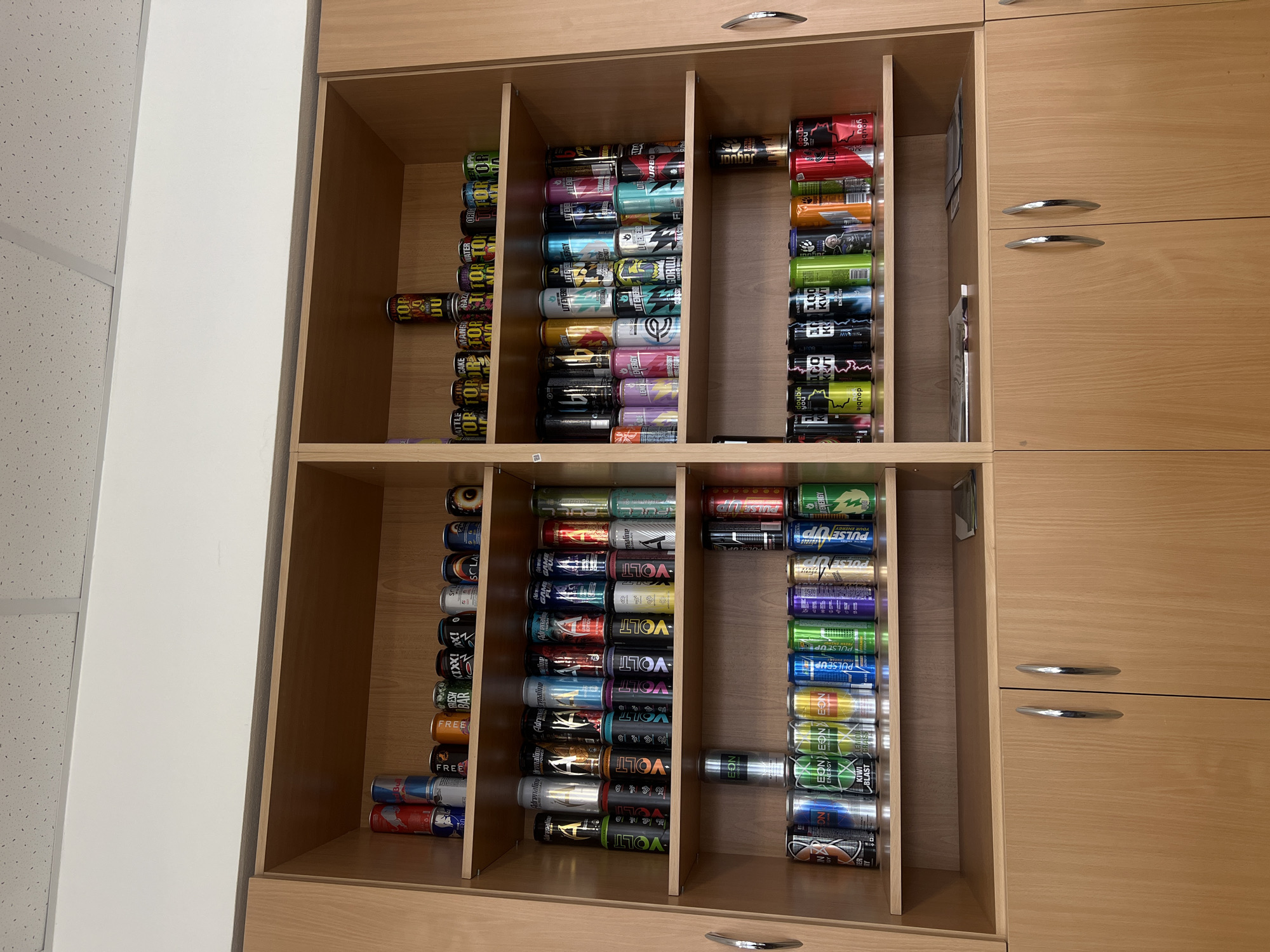 The image displays an indoor scene, likely in Chaikovsky, Russia, centered on a light-colored wooden cabinet or shelving unit filled with a large collection of energy drink cans. The cabinet features multiple shelves and vertical dividers, creating numerous compartments where the cans are neatly organized. On the right, two closed cabinet doors with silver handles are visible. To the left, a white wall section and a white, textured ceiling (possibly acoustic tiles) are present.
The primary subjects are the numerous energy drink cans, predominantly laid horizontally in stacked rows or arranged vertically. The cans exhibit a wide variety of colors, designs, and brands, suggesting a diverse collection or a comprehensive inventory.
Visible text on the cans includes brand names such as "VOLT", "TOR", "PULSEUP", "EON", "Adrenaline", "DOUBLE YOU", "JAGUAR", "LIT ENERG", "LIT TURBO", "GORILLA", "FREE", "BATTLE", "ACTIVE", "CAKE", "MANGO RAZZ", "SOLA WATER", "ORIG", "KIWI BLAST", "100 KW", and "NZ". Other discernible text includes phrases like "ENERGY", "YOUR ENERGY", "PULSE UP", "GAME", "WATER", "DU", "RUL", "BLACKOUT", and "FRESH BAR".
No people are visible, and no specific activity is taking place beyond the static display of the collection. The organized arrangement suggests a deliberate display or storage system. The setting appears to be an interior space, potentially an office, break room, or a personal collection area, under artificial lighting.
