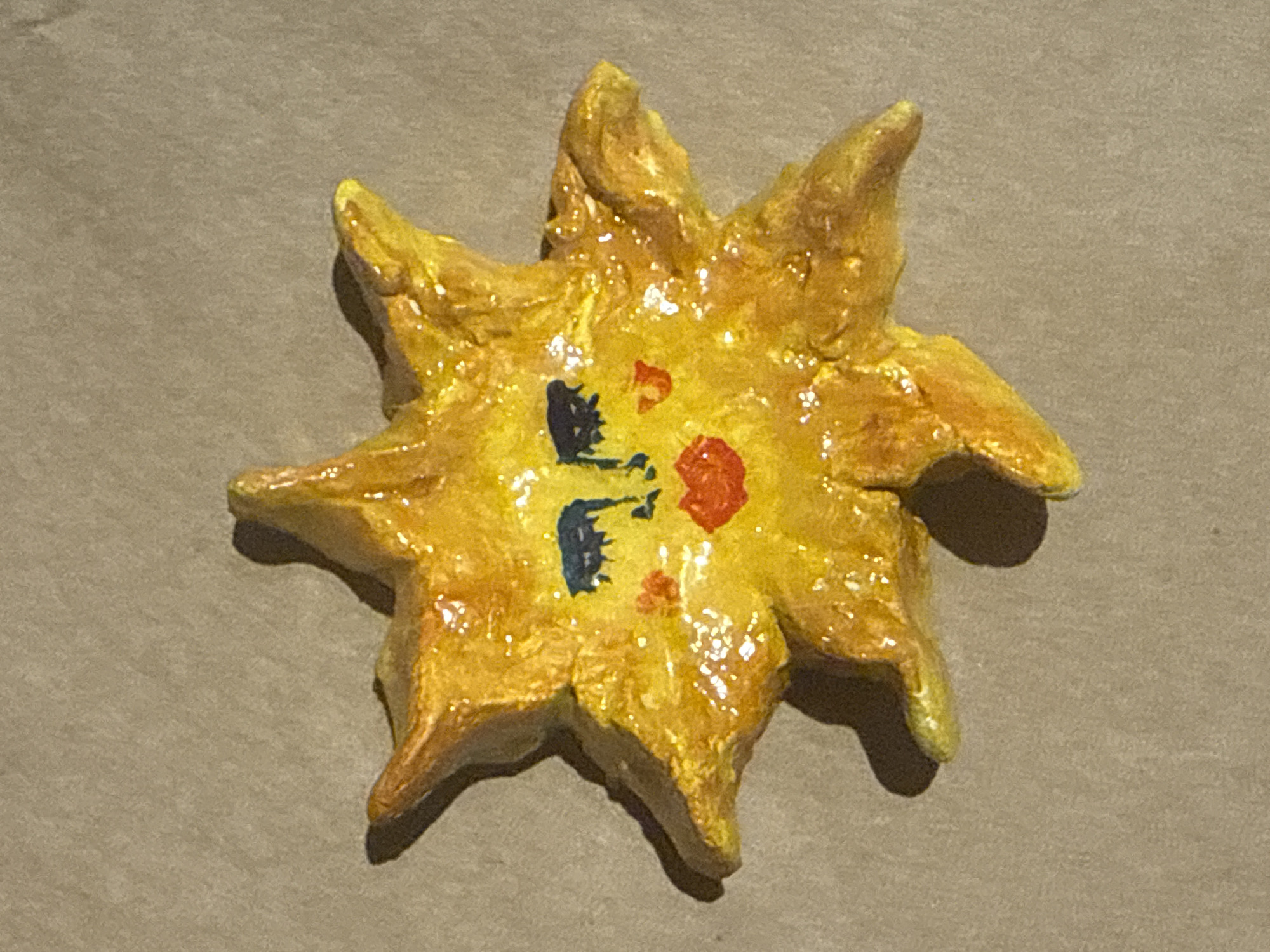 A small, handmade sun ornament is the central subject of this image, set against a neutral, textured background that resembles cardboard or craft paper. The ornament itself is crafted from a malleable material, likely clay or dough, and painted in bright, cheerful yellows with orange and red accents. It has a stylized, almost cartoonish face painted on its center, featuring blue eyes and a red mouth. The sun has eight points, or rays, radiating outward, giving it a star-like appearance. The surface of the ornament is glossy, suggesting it has been varnished or coated with a sealant, and sparkles from scattered glitter or embedded reflective material catch the light. The overall aesthetic is childlike and whimsical, indicative of a craft project. There is no visible text in the image. The lighting appears to be overhead and relatively soft, casting subtle shadows that give the ornament a three-dimensional quality. The setting is not clearly defined, but the background suggests an indoor or studio environment, possibly a craft table. The image captures a moment of stillness, focusing solely on the decorative object.
