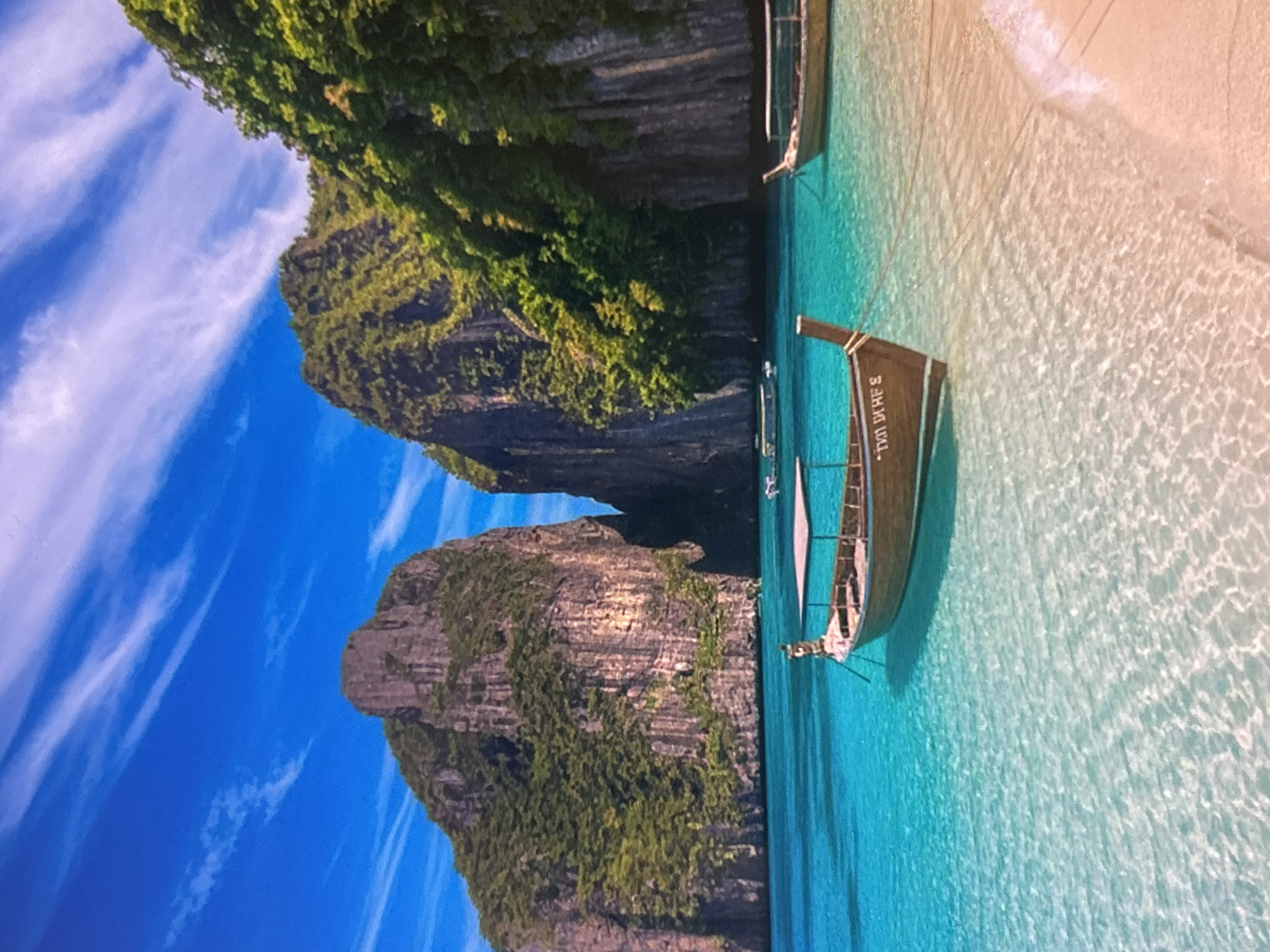 This image captures a breathtaking tropical bay, rotated 90 degrees clockwise from its natural landscape orientation. When viewed correctly, it depicts a vibrant scene featuring dramatic limestone karsts, crystal-clear turquoise waters, and traditional longtail boats under a bright blue sky.
Two towering, verdant limestone cliffs dominate the left side and background of the image, their steep faces covered in lush green vegetation. The water in the foreground and center is remarkably transparent, revealing the sandy bottom and intricate patterns of light reflecting on the surface. To the right, a pristine sandy beach curves gently into the calm waters.
Several longtail boats are visible in the bay. One prominent boat, dark-hulled with a light-colored canopy or sail structure, is positioned in the middle ground, slightly to the right of the center. On its side, faded text, which appears to be Thai script, is visible. Other boats are smaller and further in the background, hinting at more activity beyond the immediate foreground.
The sky is a brilliant blue with soft, wispy white clouds streaking across it, suggesting clear and sunny weather. The overall impression is one of a tranquil and idyllic tropical paradise, likely a popular tourist destination in Southeast Asia, such as Thailand, known for its distinctive karst formations and longtail boats. There are no people explicitly visible, emphasizing the natural beauty and serene atmosphere of the location. The time of day appears to be bright daylight, possibly late morning or early afternoon.