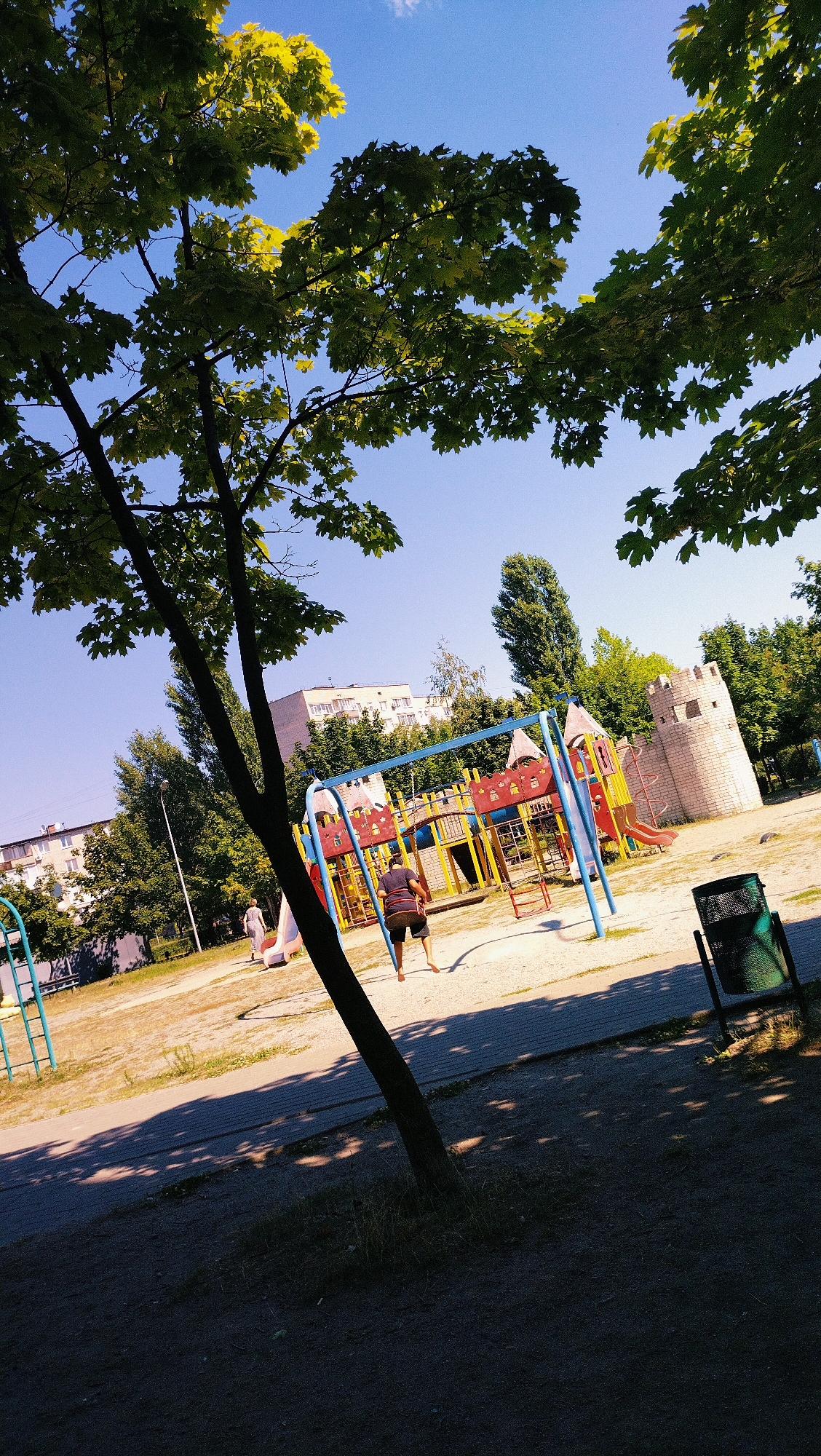 An outdoor, low-angle, eye-level shot captures a children's playground on a sunny day. The foreground is dominated by a dark, tree-lined area, casting dappled shadows across the ground. In the midground, a colorful playground structure with slides and climbing elements is visible, featuring a beige stone castle-like tower on the right. A person is on a swing, their legs visible as they propel themselves. Another person walks in the background towards a slide. Beyond the playground, residential buildings and tall green trees form the backdrop under a clear blue sky. A dark green trash can stands near the right edge of the frame. The overall environment appears to be a public park or recreational area within a city.