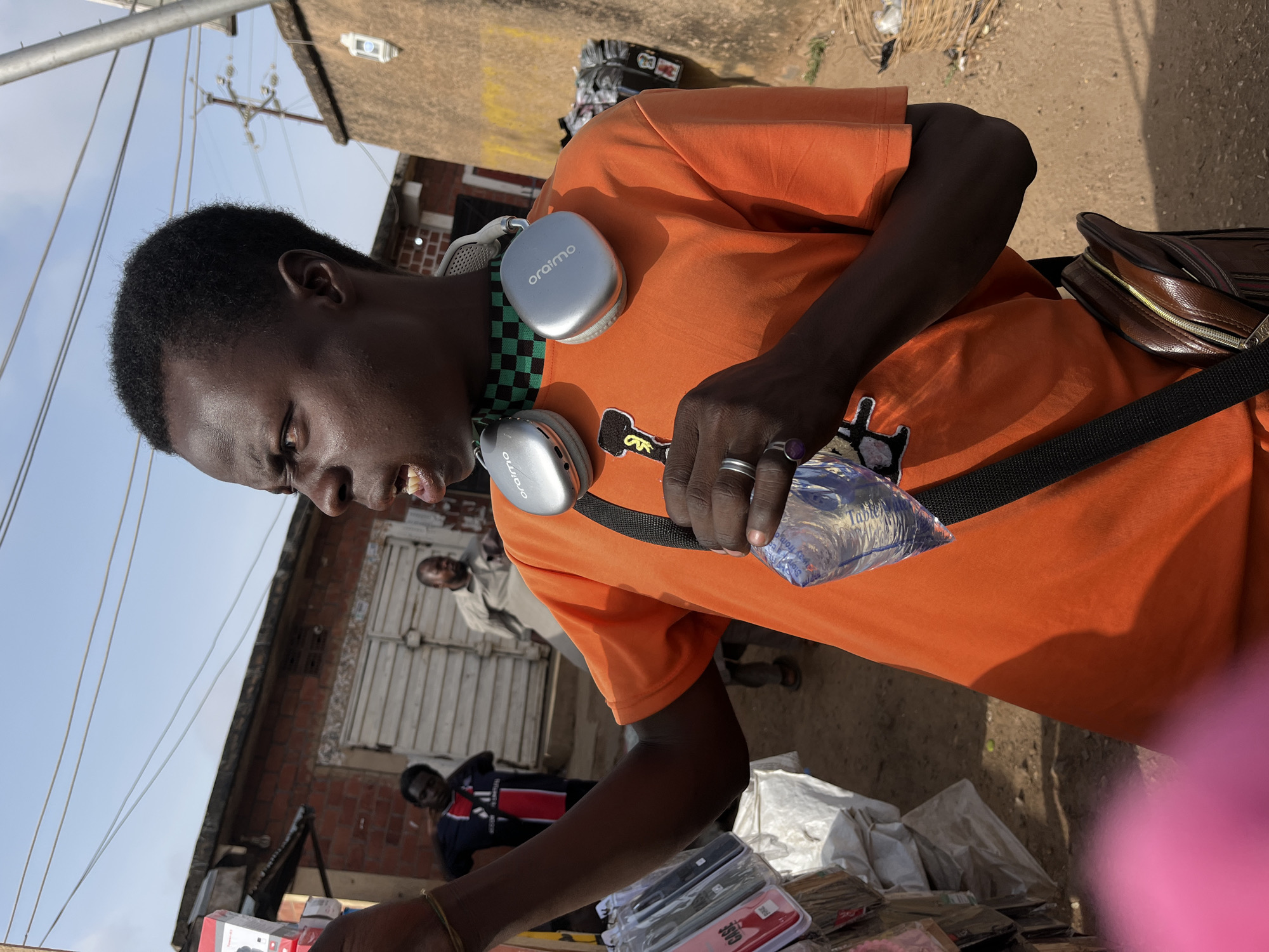 An individual stands in Gura Top, Nigeria, wearing an orange shirt, black strap across the torso, and silver "oraimo" headphones. They hold a plastic bag and wear rings on their fingers. In the background are buildings constructed with orange bricks and gray steel garage doors, an individual wearing a blue and red shirt, and white bags. The ground appears to be a dirt surface.