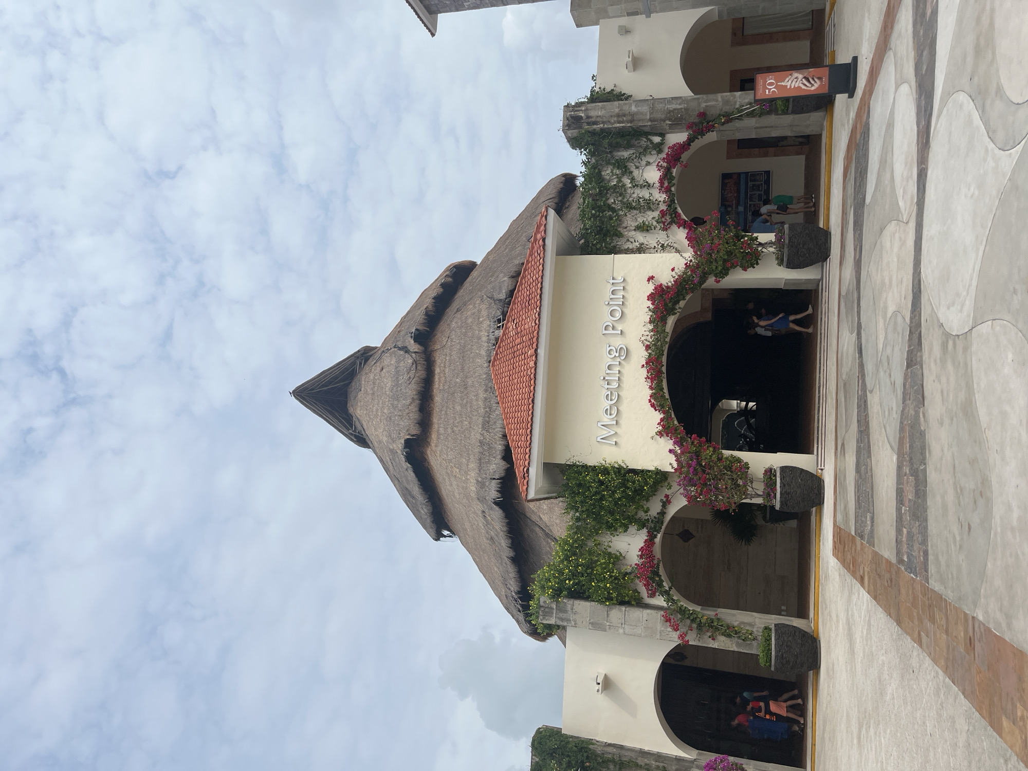 The image shows a building in Playa del Carmen, Mexico, with a thatched roof and a sign that reads "Meeting Point." The building is surrounded by flowering vines and plants. People can be seen entering and exiting the building. The ground is paved with a decorative pattern. The sky is cloudy.